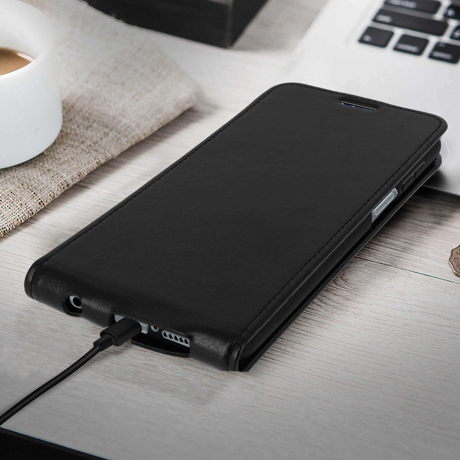 Un téléphone noir dans un étui en cuir noir, en charge sur une table en bois.