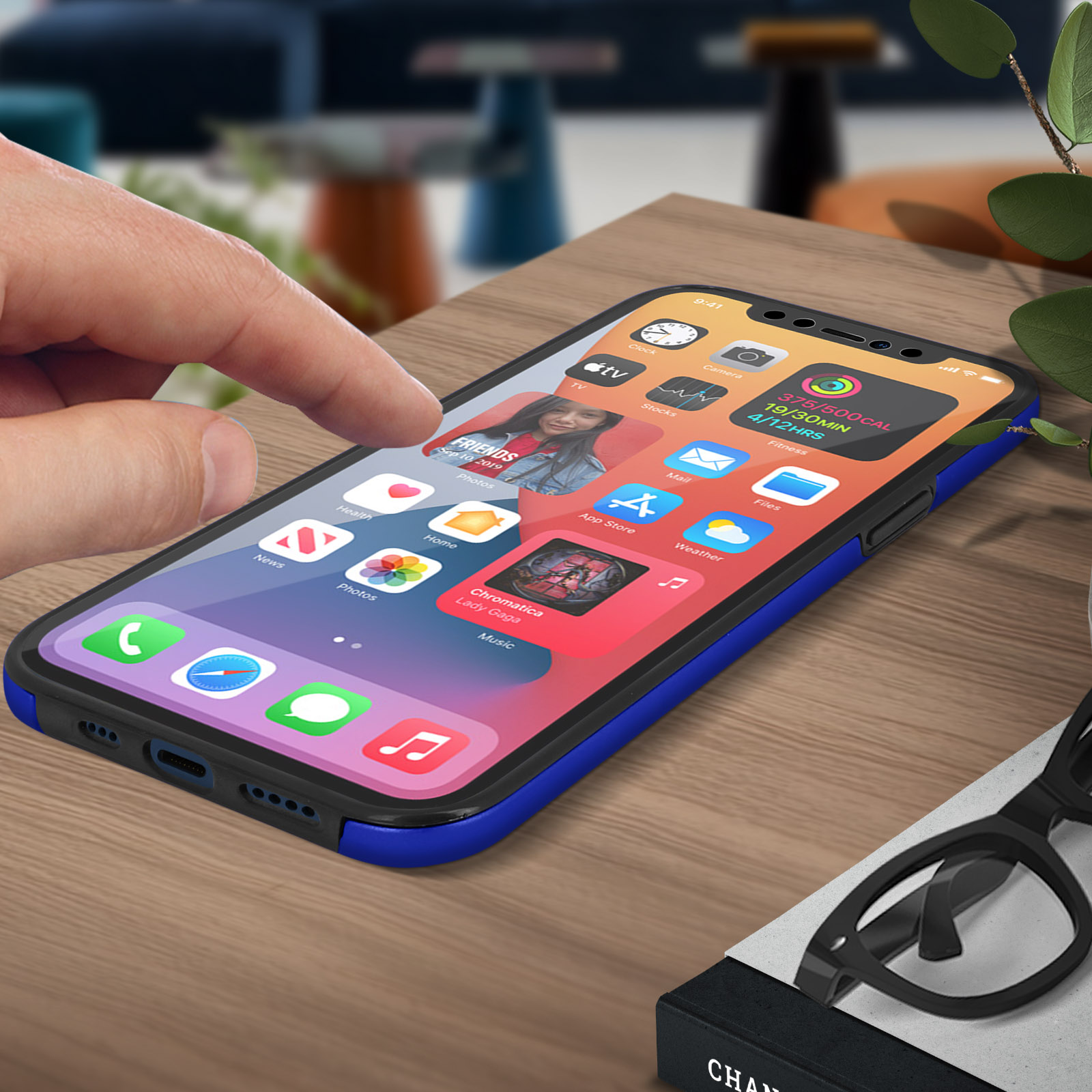Main touchant l'écran d'un smartphone avec coque bleue, sur une table en bois. Une paire de lunettes.