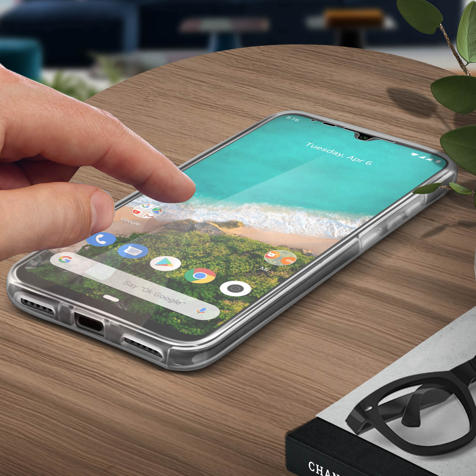 Een hand die het telefoonscherm aanraakt. De telefoon staat op een houten tafel. Bril en planten.