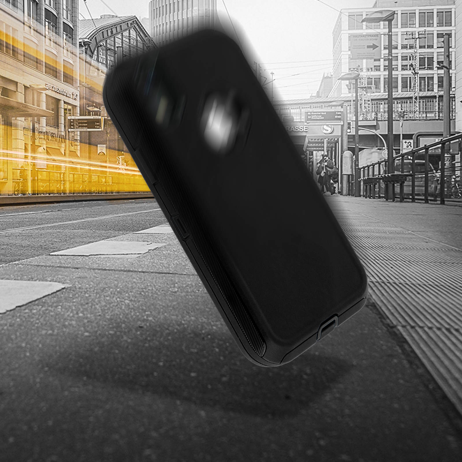 Una custodia nera per telefono che cade in aria su uno sfondo sfocato di strada cittadina in scala di grigi.