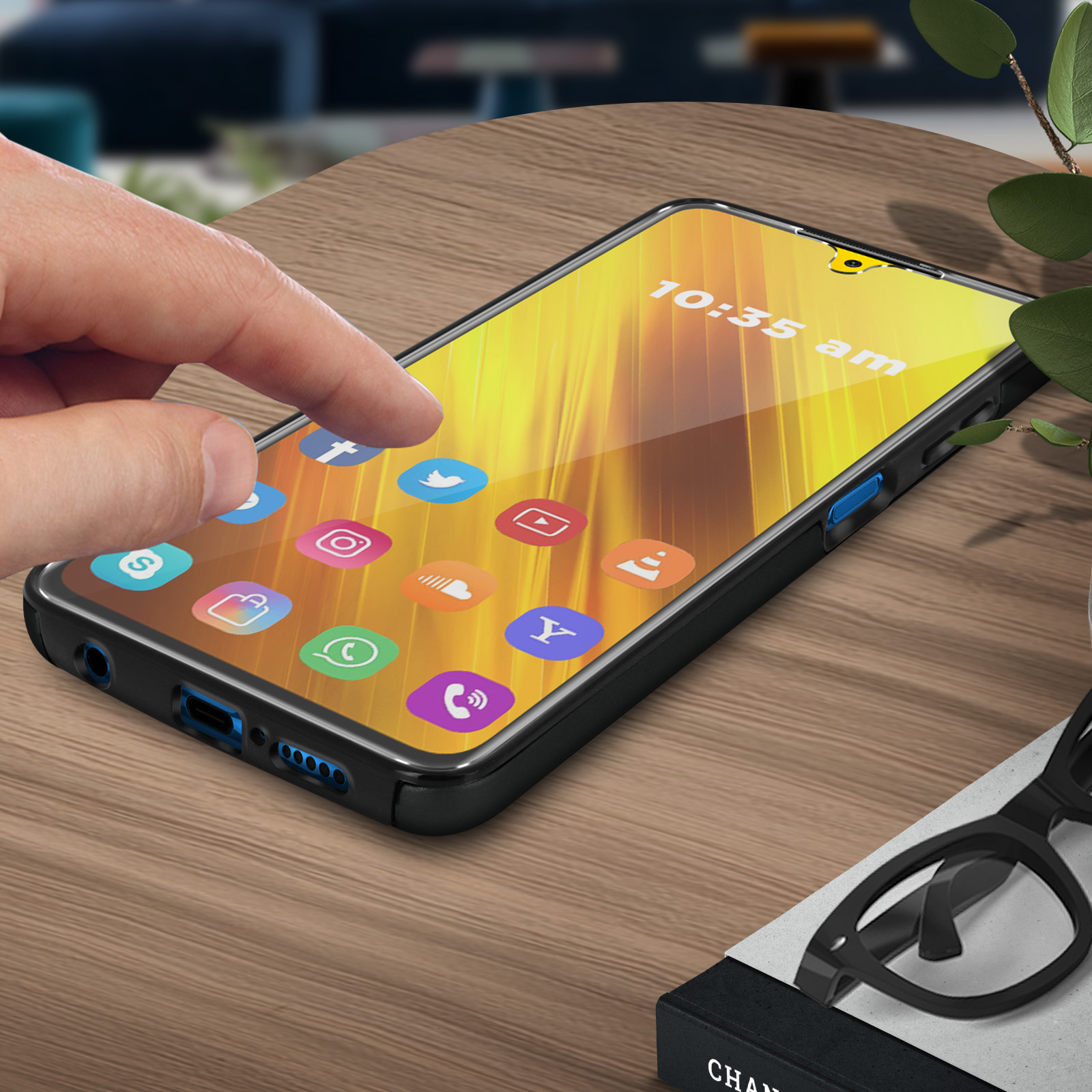 Una mano tocca lo schermo del telefono con le app. Il telefono è su un tavolo di legno, occhiali e pianta sullo sfondo.
