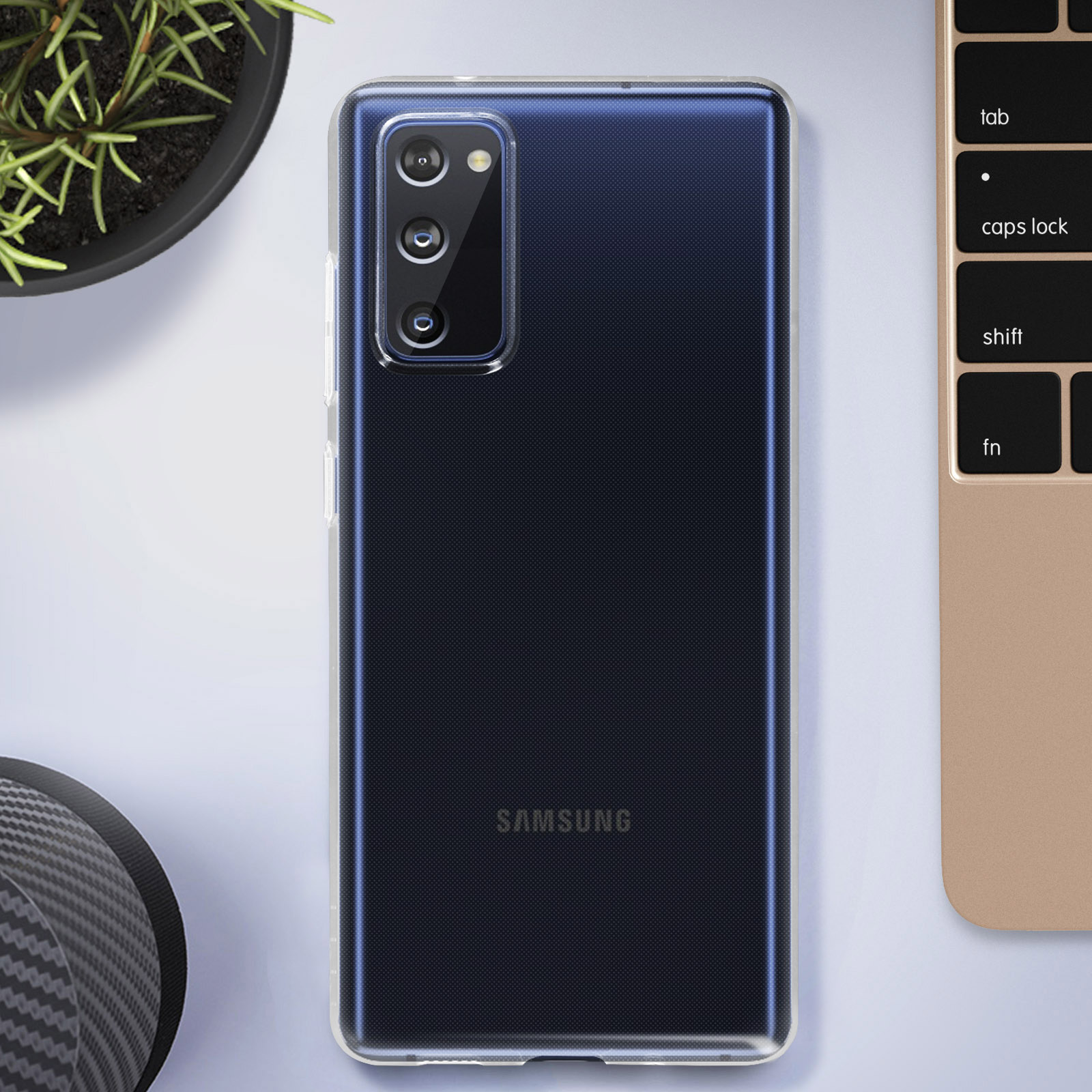 Telefono Samsung in custodia trasparente, su una scrivania vicino a una tastiera di un laptop e una pianta in vaso.