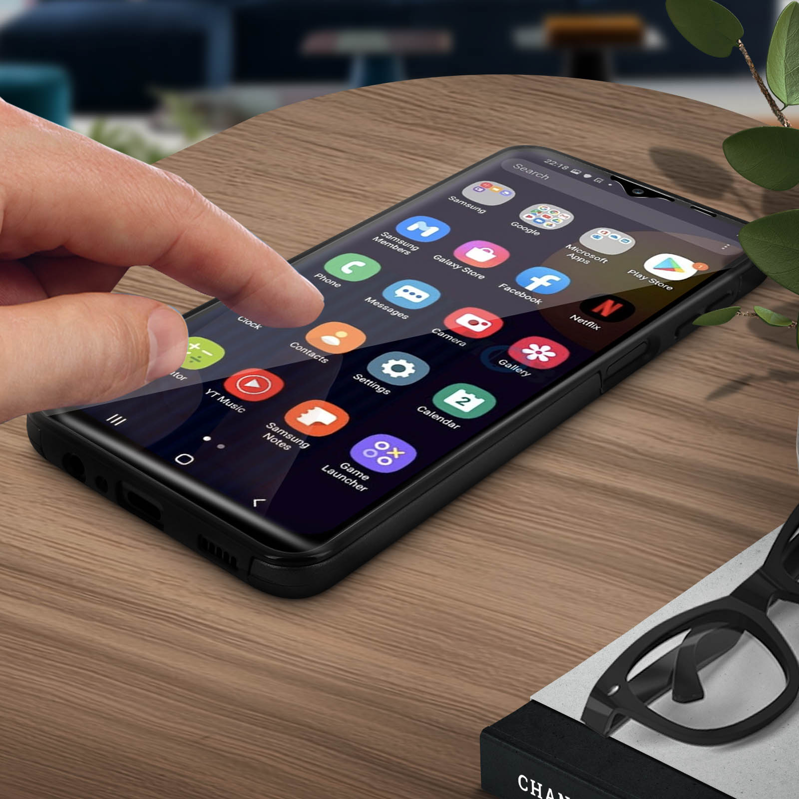 Een hand raakt een telefoonscherm aan met app-pictogrammen. Een kopje, bril en pen liggen ook op tafel.