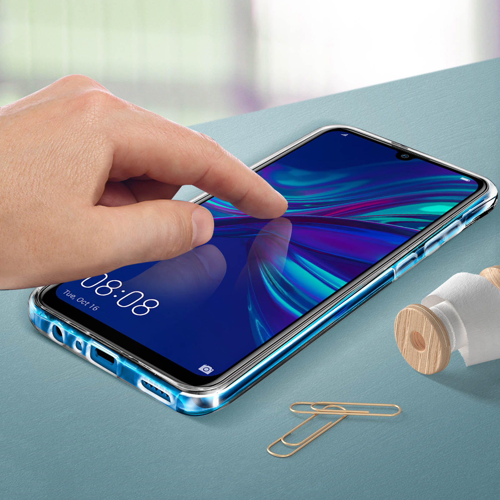 Une main touche un téléphone. L'écran affiche une image colorée. Il y a des trombones sur la table.