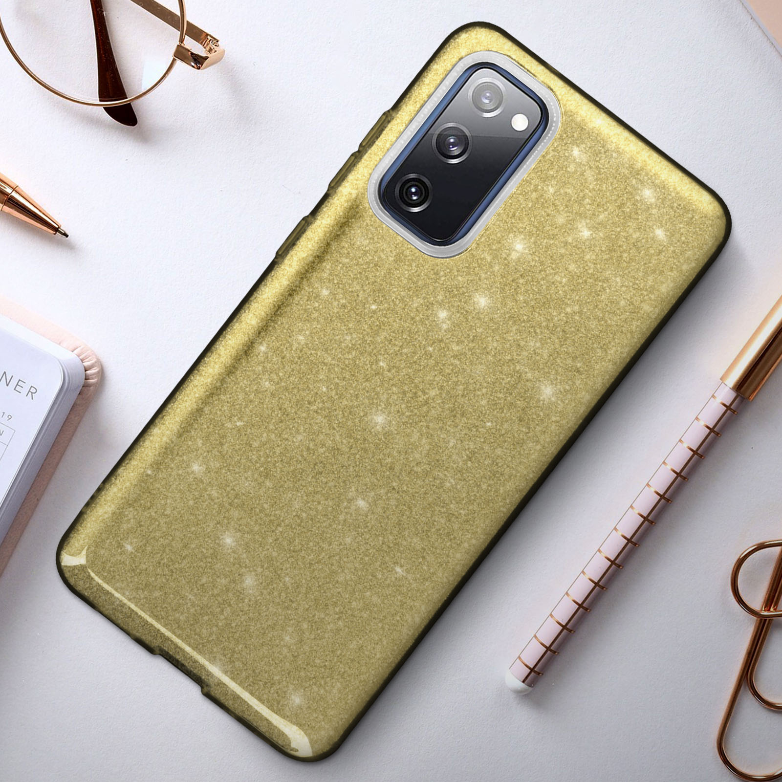 Custodia per telefono con glitter dorati su una scrivania con penna, occhiali e blocco note. La custodia ha un bordo trasparente.