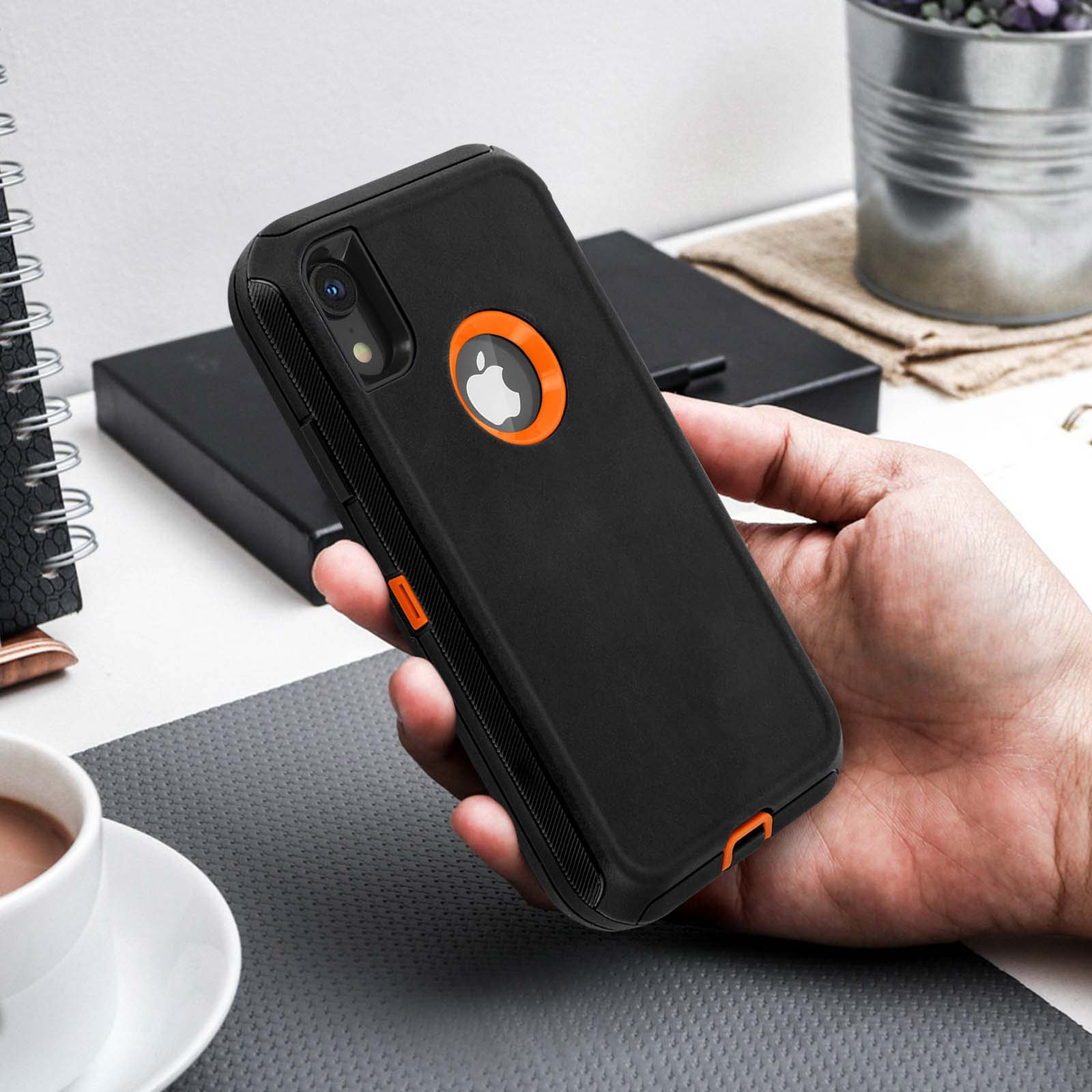 Mano che tiene una custodia nera per telefono con dettagli arancioni. Tazza bianca e superficie grigia sullo sfondo.