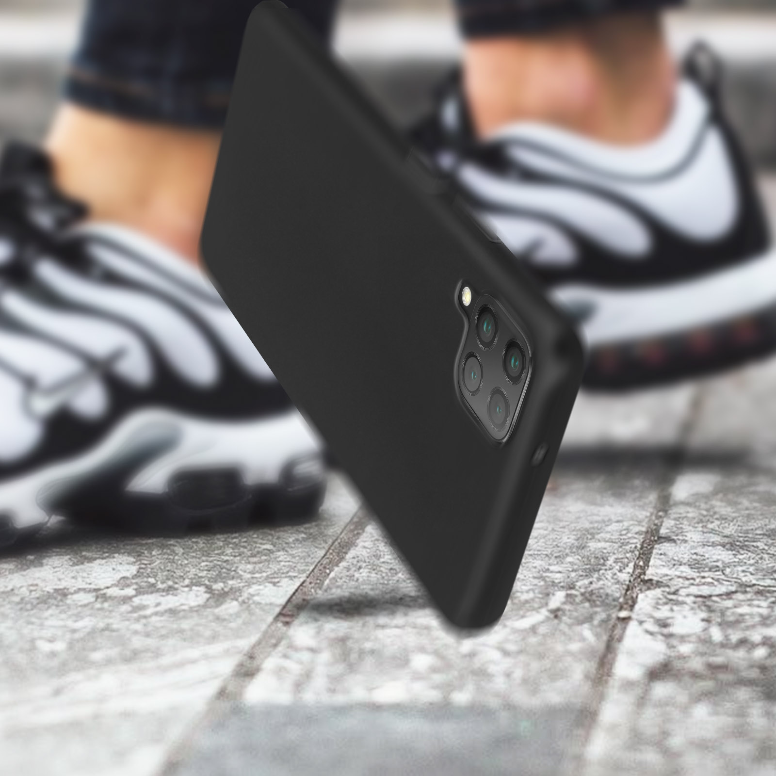 Een zwarte telefoonhoes op een gestructureerd oppervlak met een persoon met sneakers op de achtergrond.
