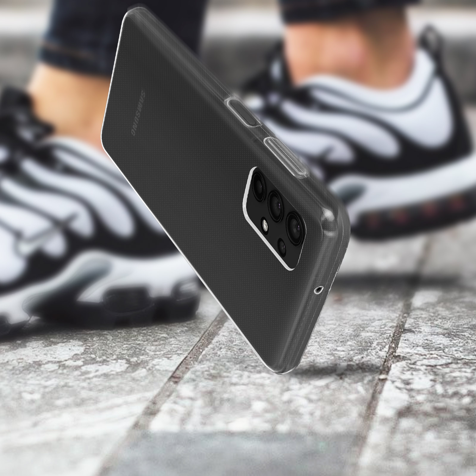 Een telefoon in een zwarte hoes valt op een betonnen oppervlak. De achtergrond toont vage benen en schoenen.
