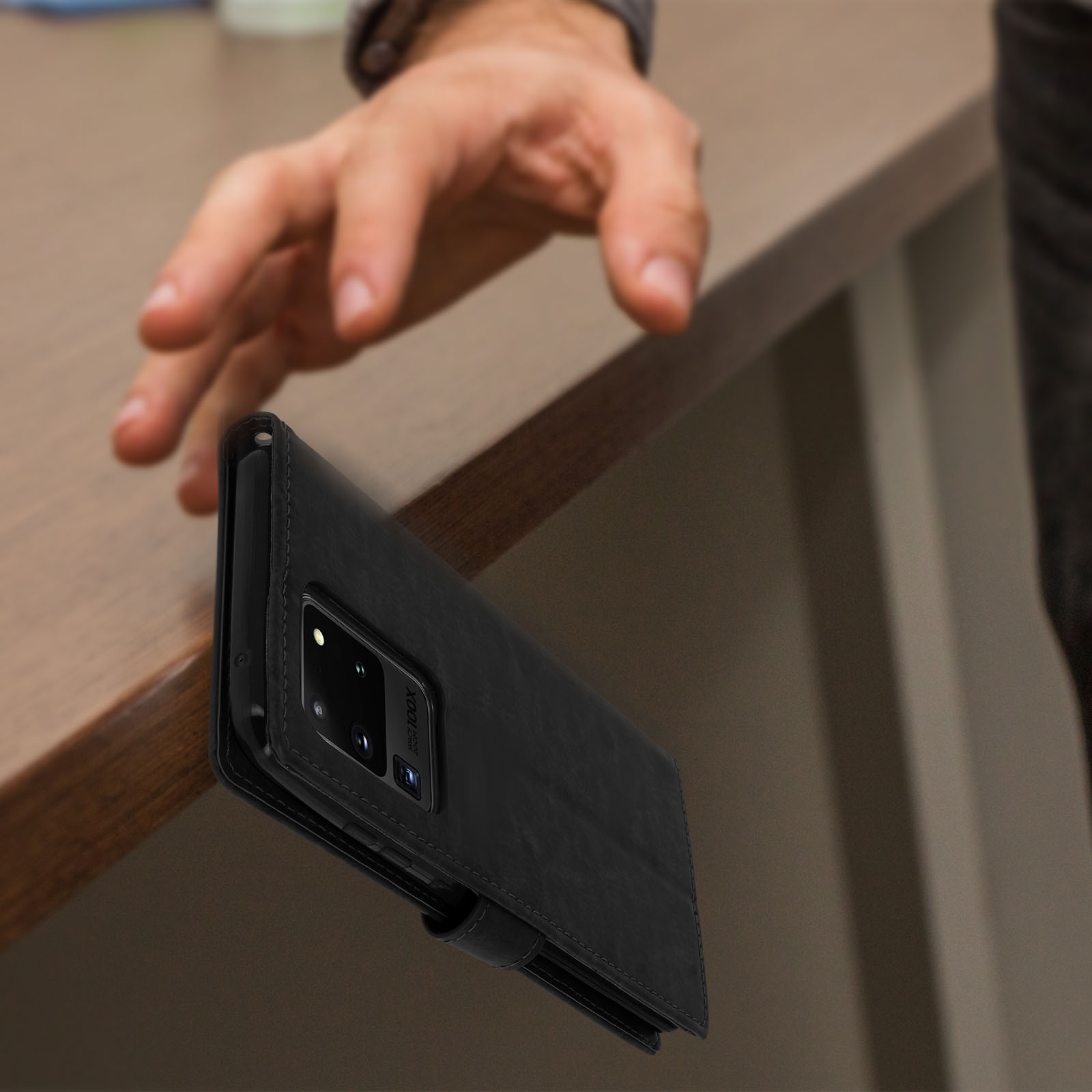 Een zwarte telefoonhoes op een bruine tafel. Een hand reikt naar de hoes.