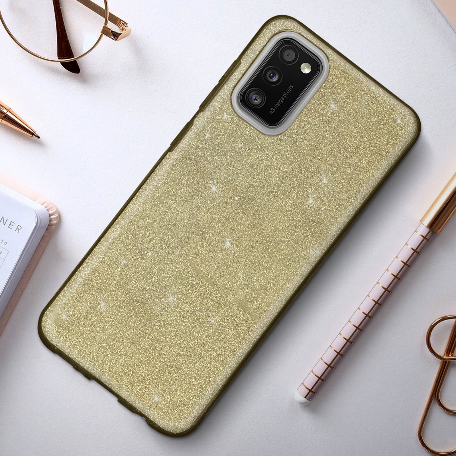 Een telefoonhoes met een glinsterende gouden afwerking ligt op een tafel met een bril, pen en notitieblok.