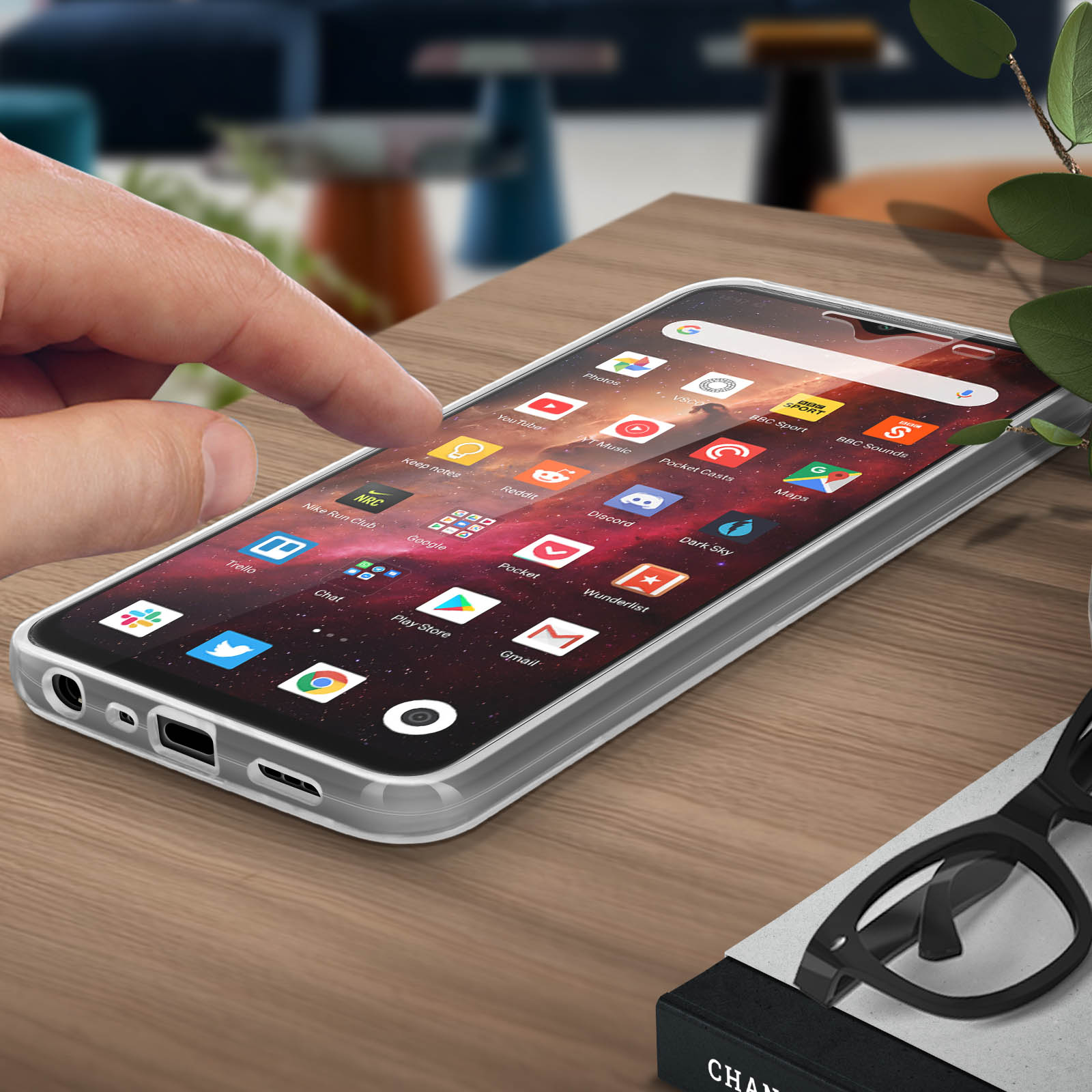 Main touchant l'écran du téléphone affichant des icônes d'applications. Téléphone dans une coque transparente, sur un bureau avec des lunettes et une plante.