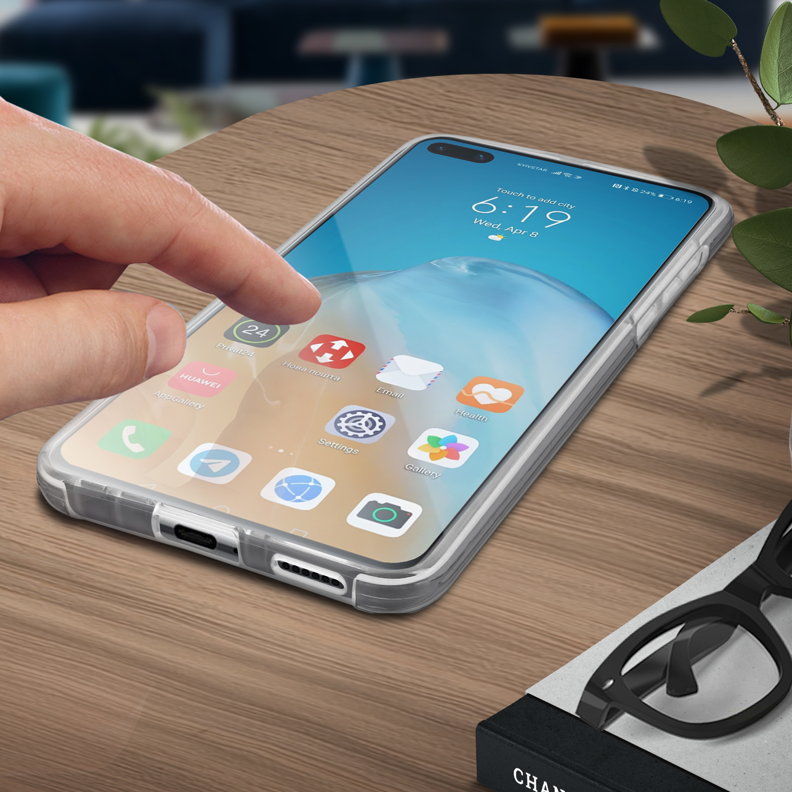 Een hand raakt een telefoonscherm met apps aan. Telefoon in een doorzichtige hoes op een houten tafel. Bril en plant in de buurt.