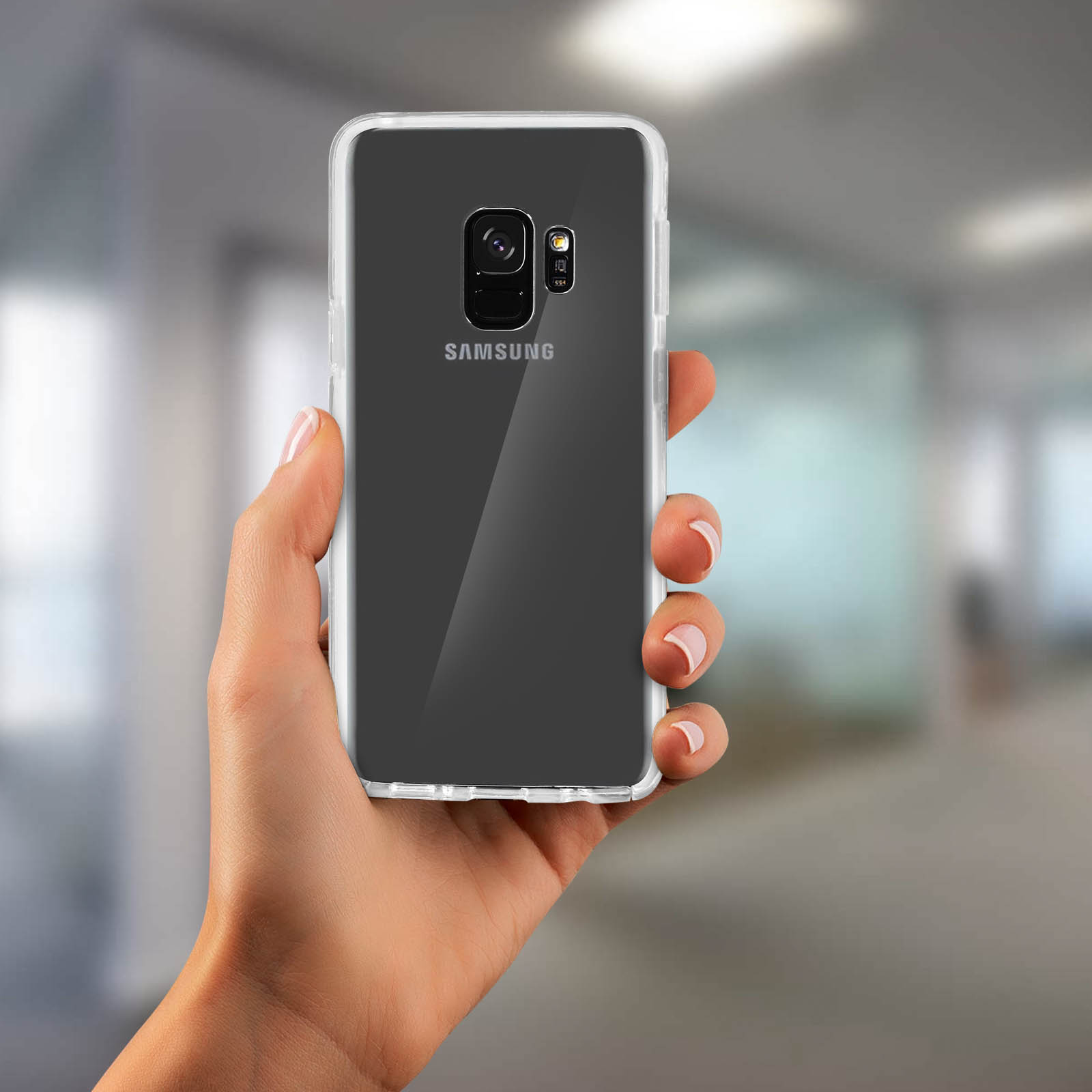Mano che tiene un telefono con una custodia trasparente, logo SAMSUNG sul retro. Lo sfondo è sfocato.