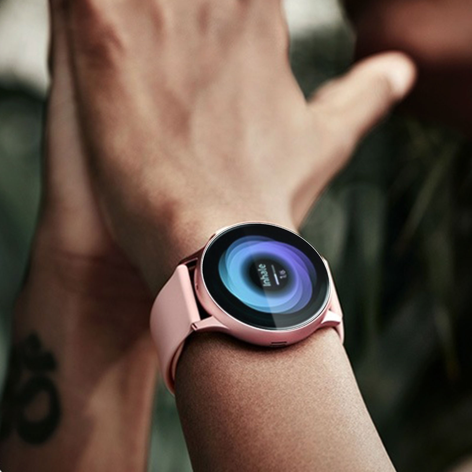 Polso di una persona con uno smartwatch rosa. Il quadrante mostra 'Inhale'. La pelle ha un tatuaggio.