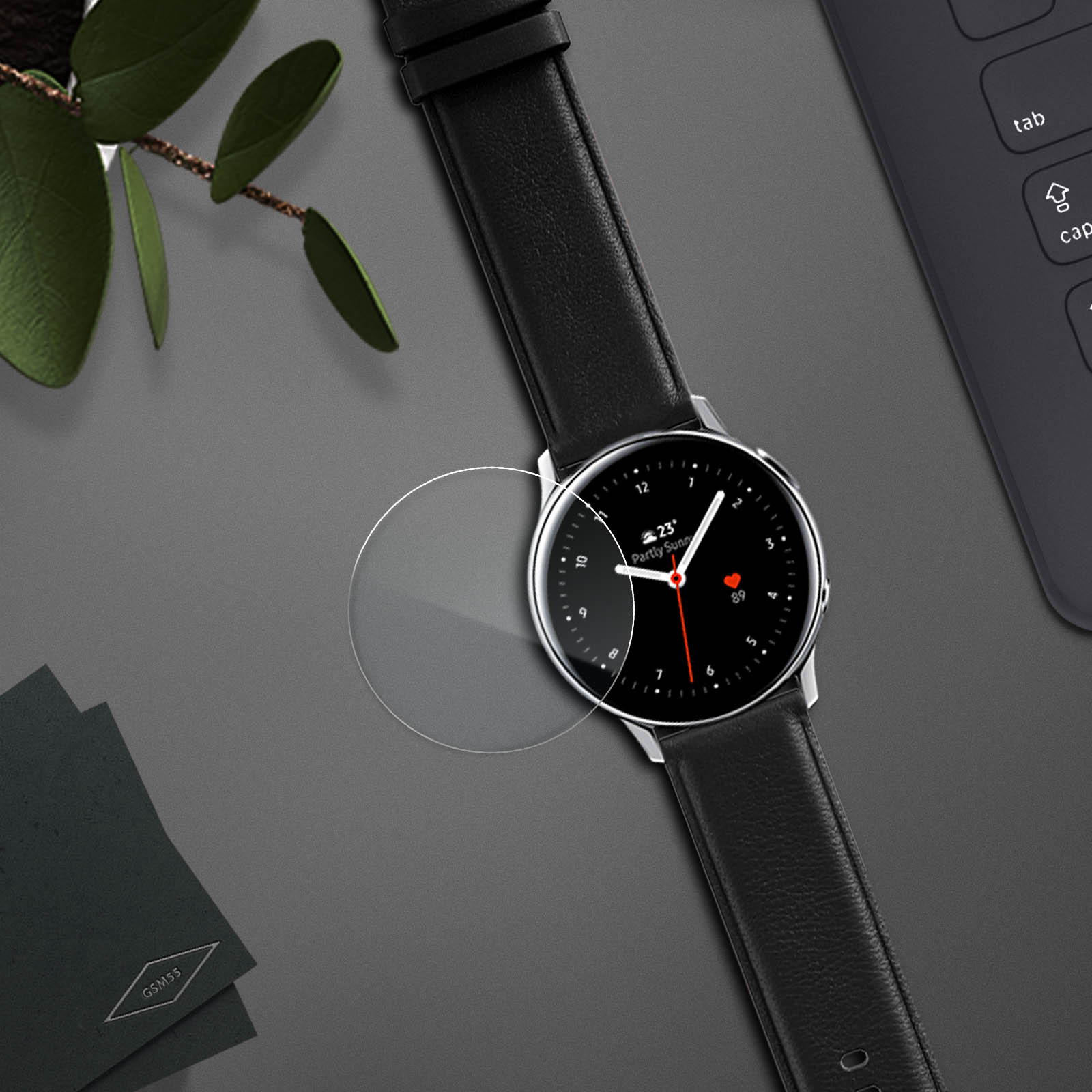 Uno smartwatch con cinturino in pelle nera, protezione schermo e quadrante nero. Tastiera e foglie sullo sfondo.