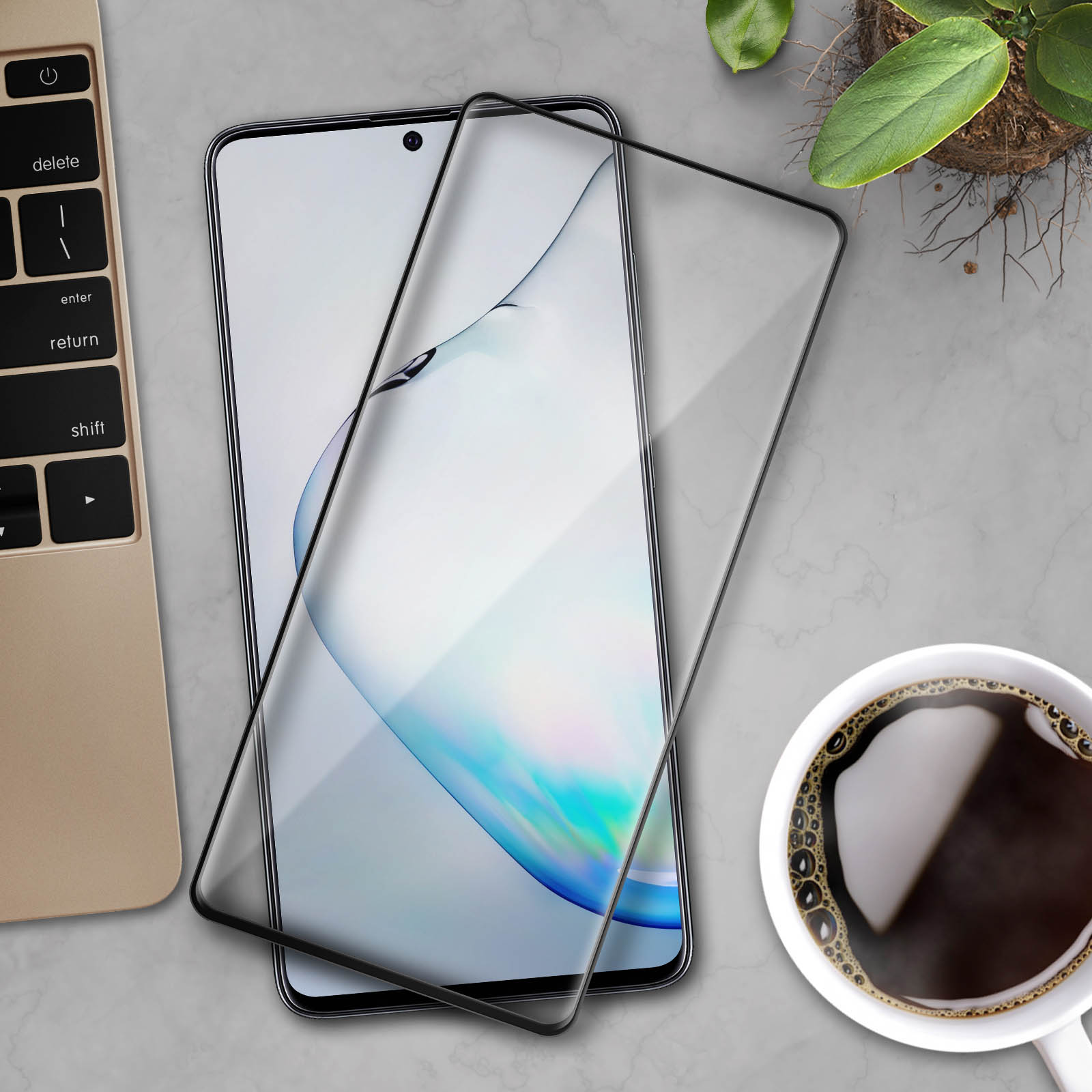Uno smartphone con una protezione per lo schermo su un tavolo. Una tazza di caffè, una tastiera e una piccola pianta sono visibili.