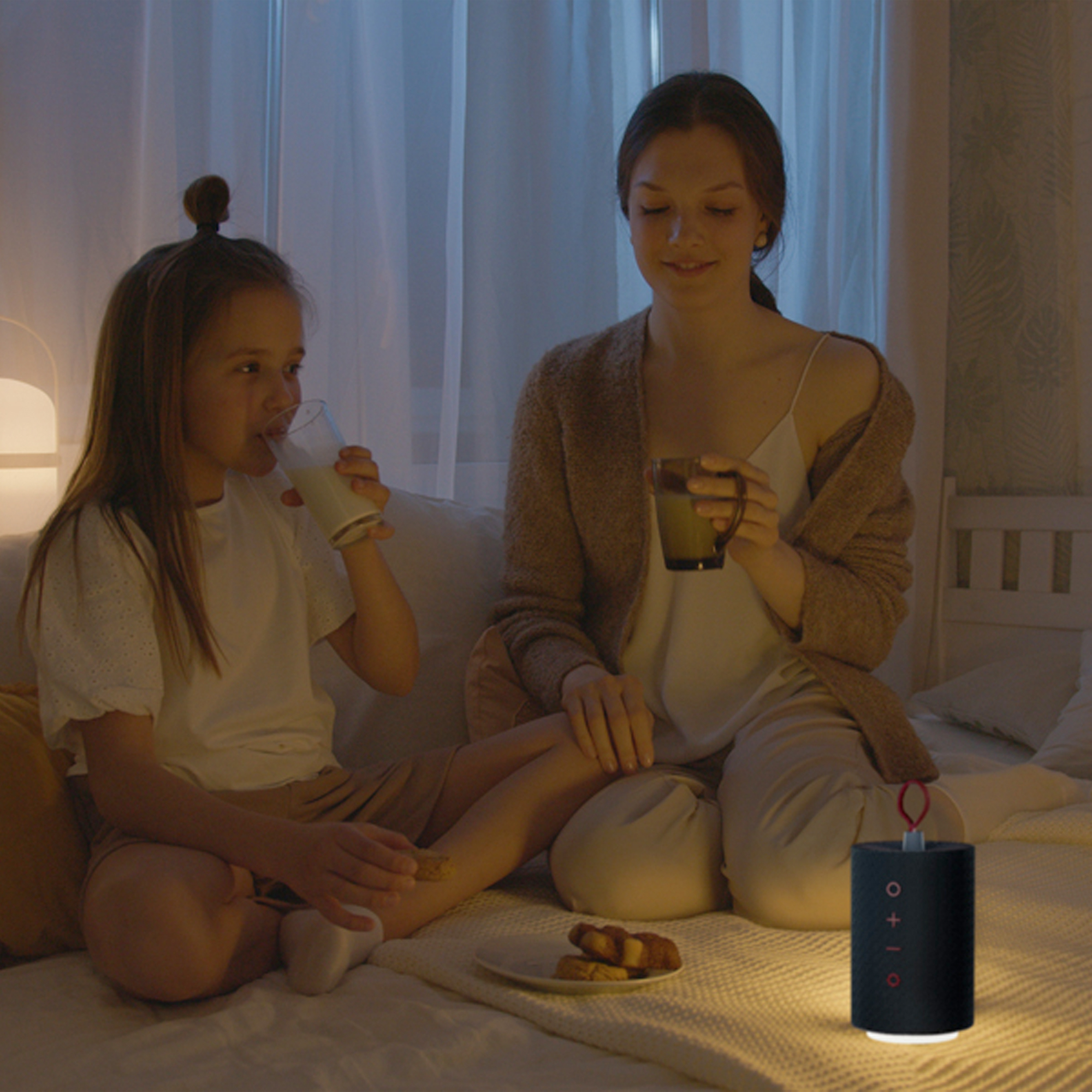 Mutter und Tochter sitzen auf einem Bett und trinken aus Tassen, mit einem Lautsprecher daneben.