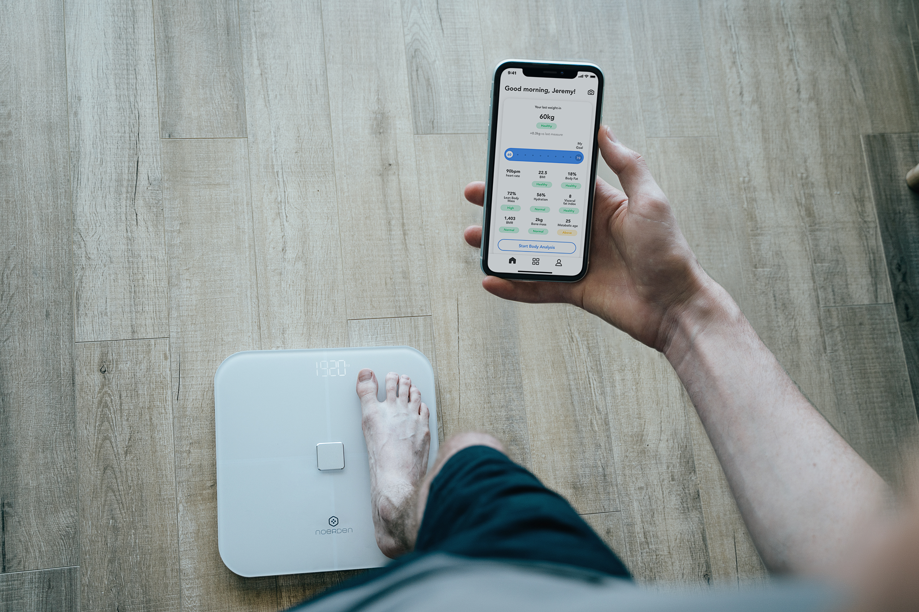 Eine Person steht auf einer Waage und hält ein Telefon mit Gewichtsdaten. Ein Kind und eine weitere Waage im Hintergrund.