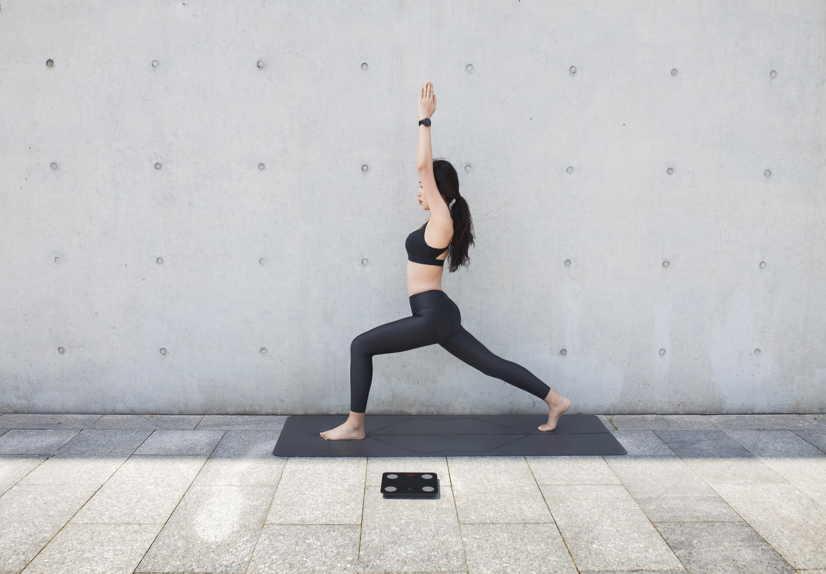 Eine Frau macht Yoga. Sie ist in einer Kriegerpose mit erhobenen Armen. Eine Waage liegt zu ihren Füßen.