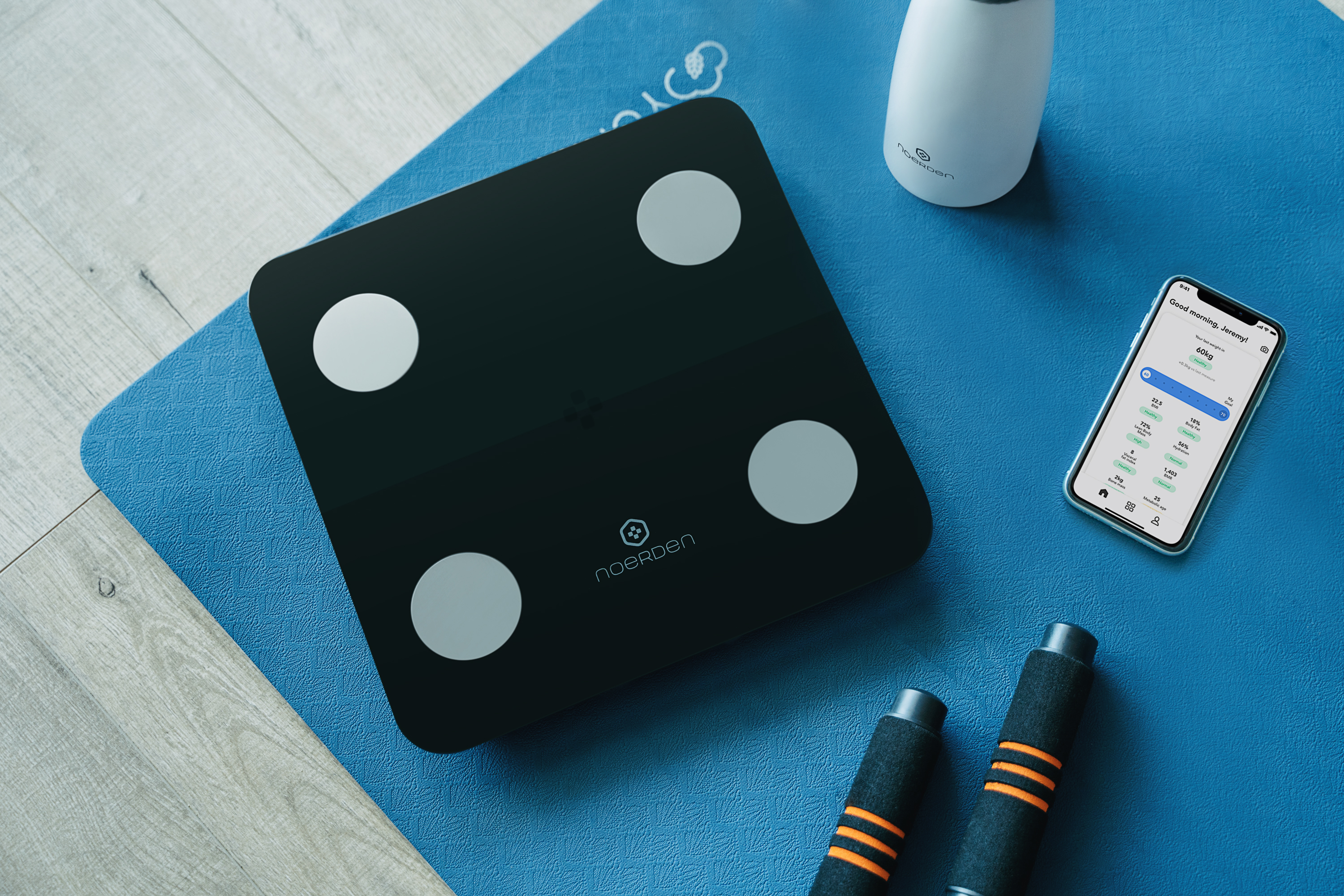 Schwarze Smart-Waage auf blauer Matte mit Telefon und Zubehör. Marke Noerden sichtbar.