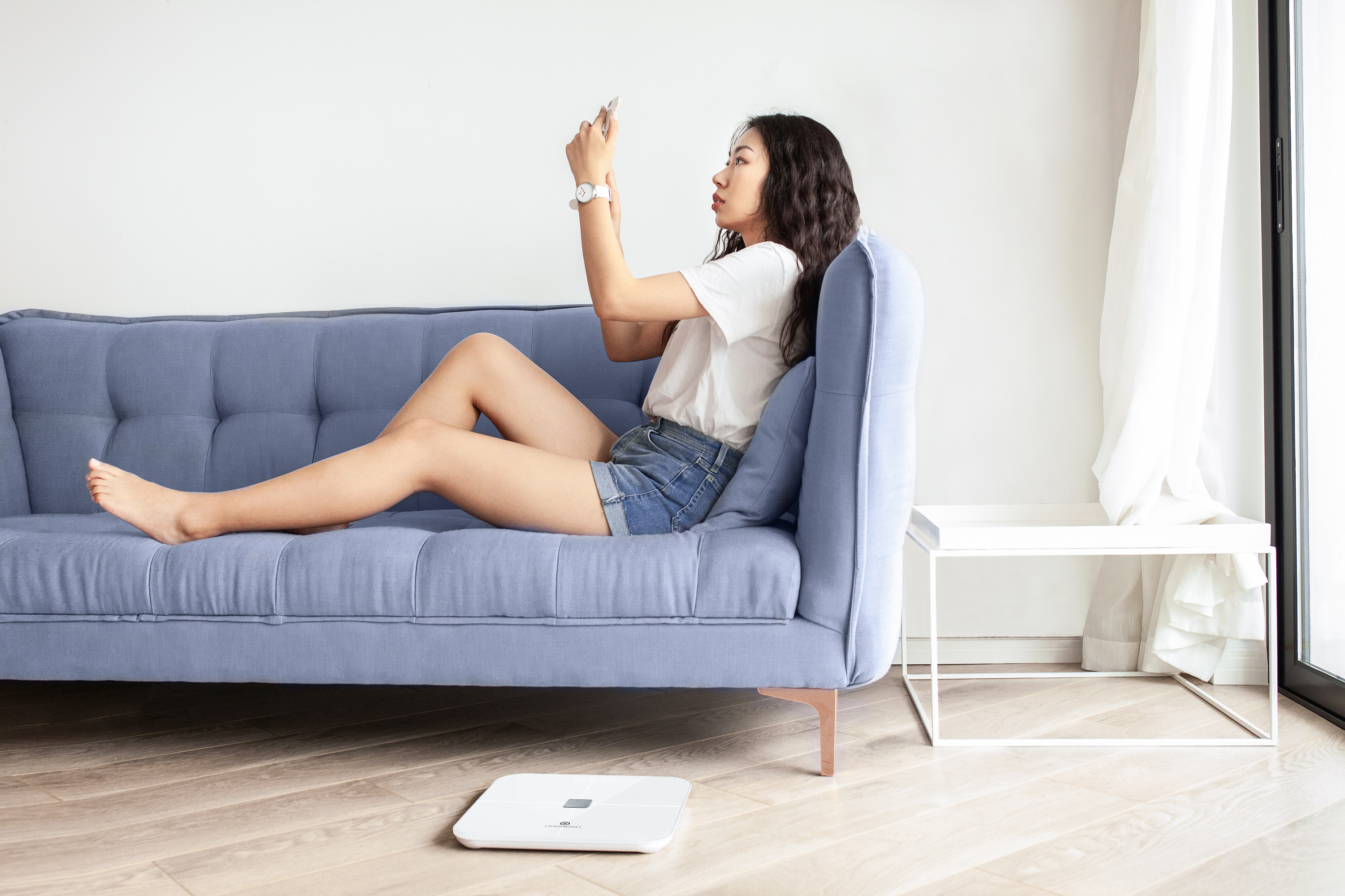Eine Frau sitzt auf einem blauen Sofa und benutzt ein Telefon, eine weiße Waage liegt auf dem Boden.
