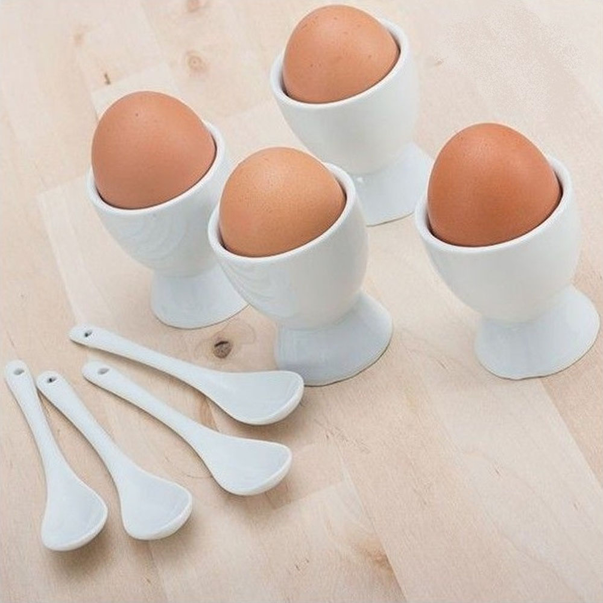 Quattro uova in portauova bianchi, con cucchiai su una superficie di legno. Le uova sono marroni e i cucchiai bianchi.