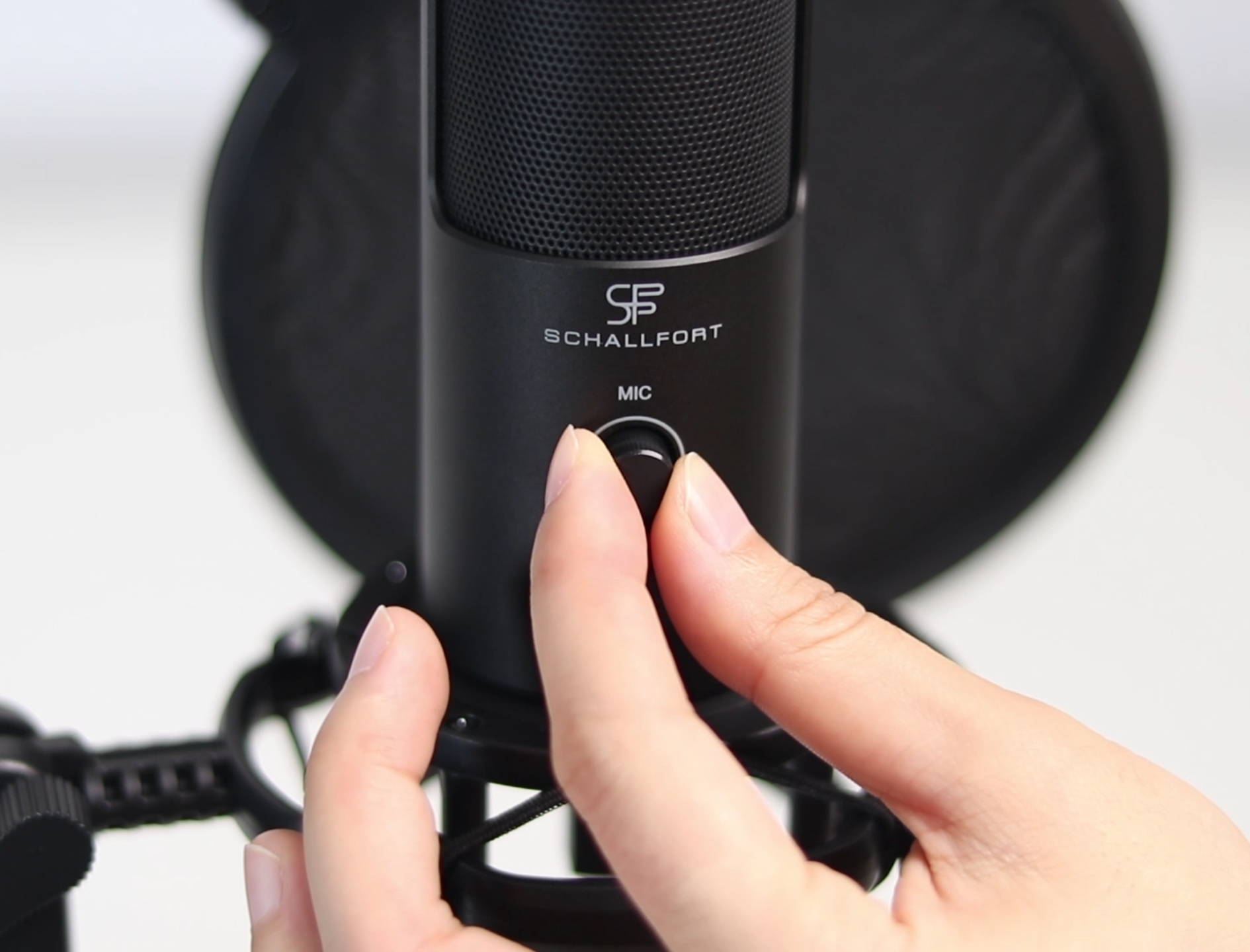 Ein schwarzes Mikrofon mit Pop-Filter. Eine Hand stellt einen Drehknopf am Mikrofon mit der Aufschrift 'MIC' ein.