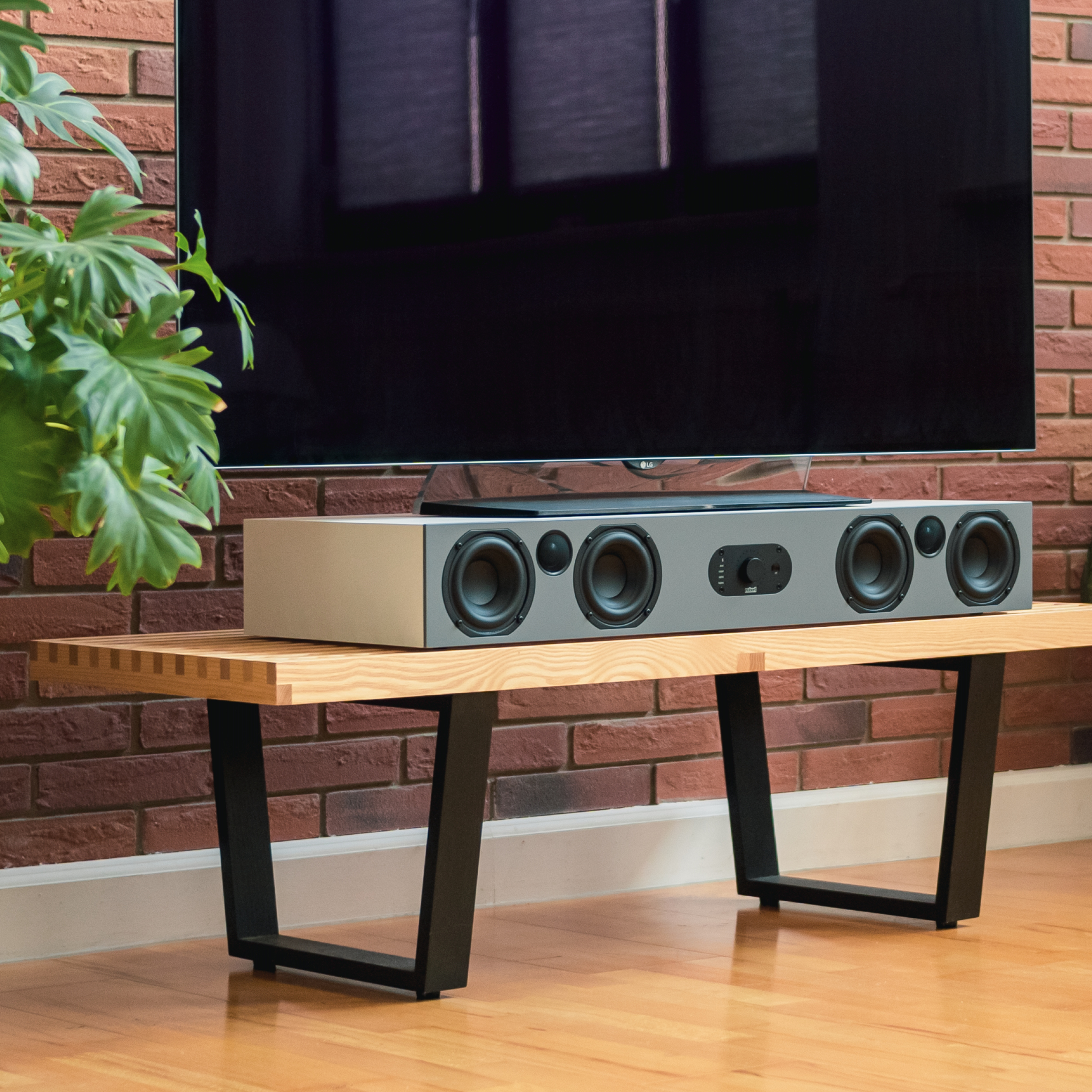 Graue Soundbar auf einer Holzbank vor einer Backsteinmauer und einem Fernseher, mit einer Pflanze in der Nähe.