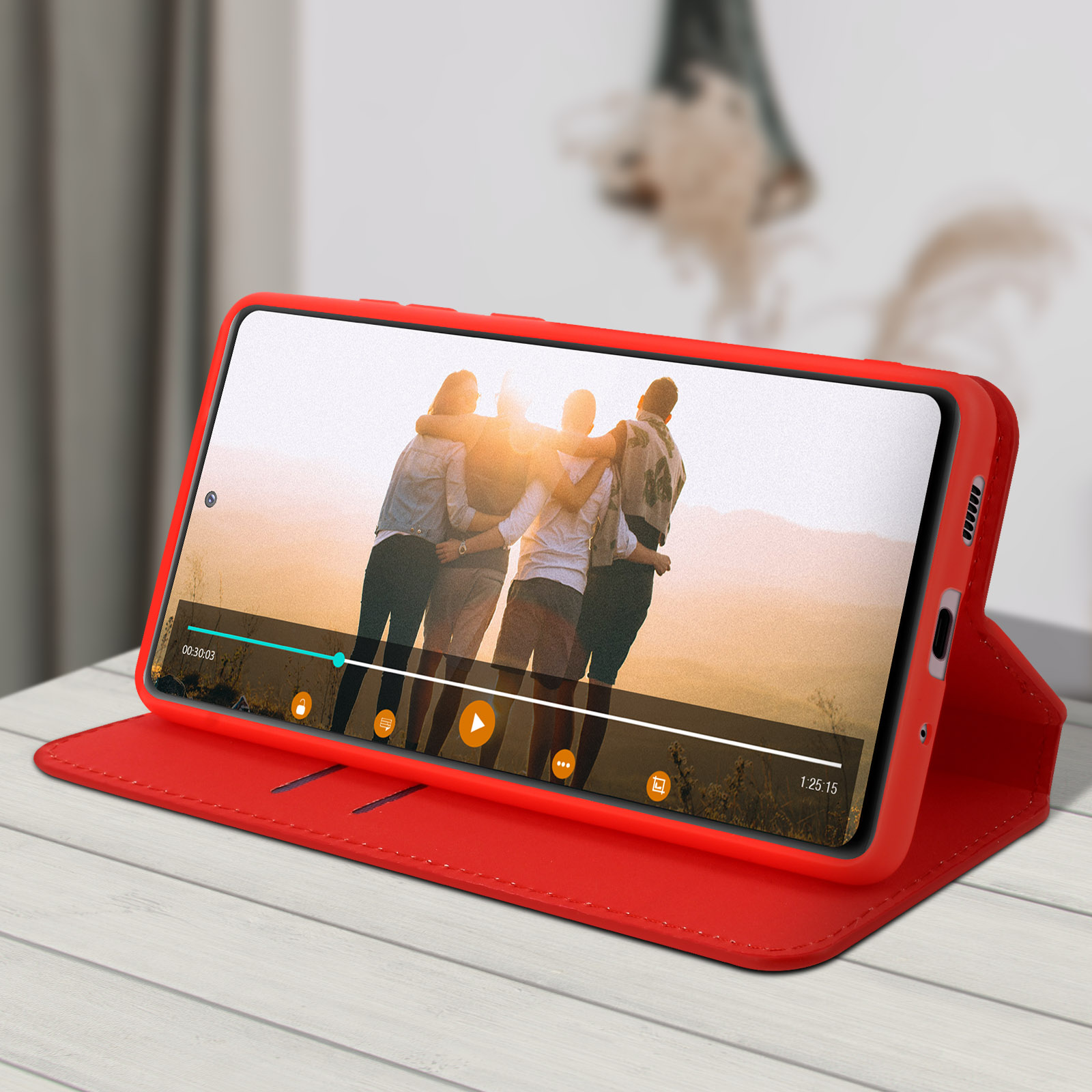 Rote Handyhülle mit geöffnetem Telefon, das ein Video von Menschen zeigt. Weißer Holztisch, unscharfer Hintergrund.