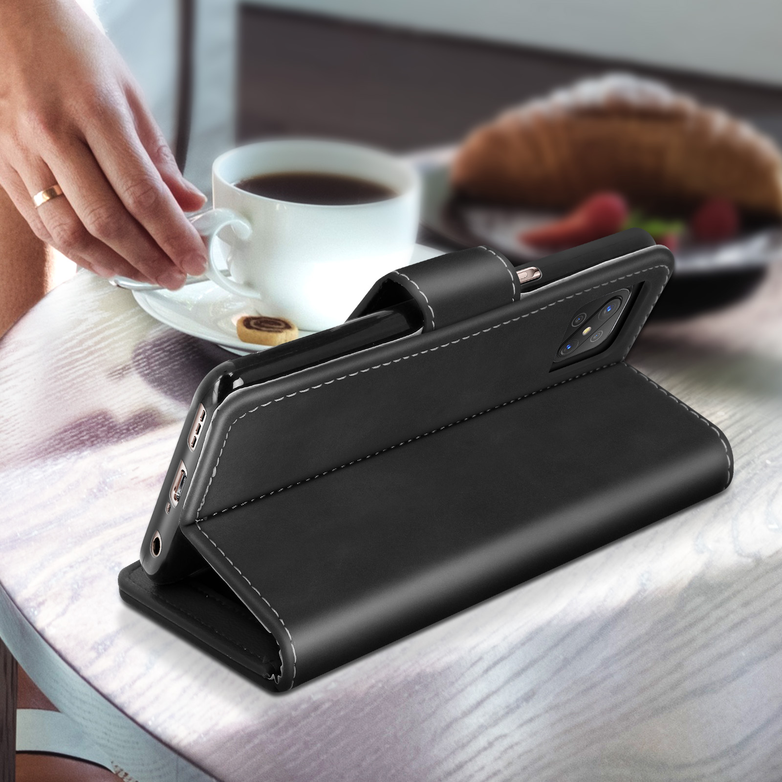 Custodia nera per telefono su un tavolo, il telefono è in mostra. Caffè, mano e croissant presenti.