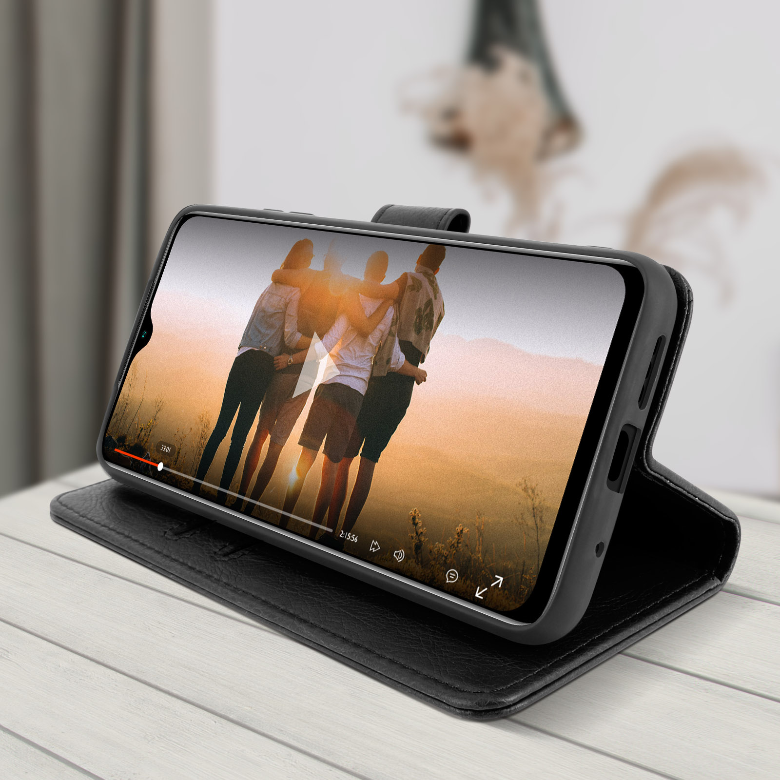 Un téléphone affiche une vidéo de trois personnes, posé sur un étui noir, sur une table en bois.