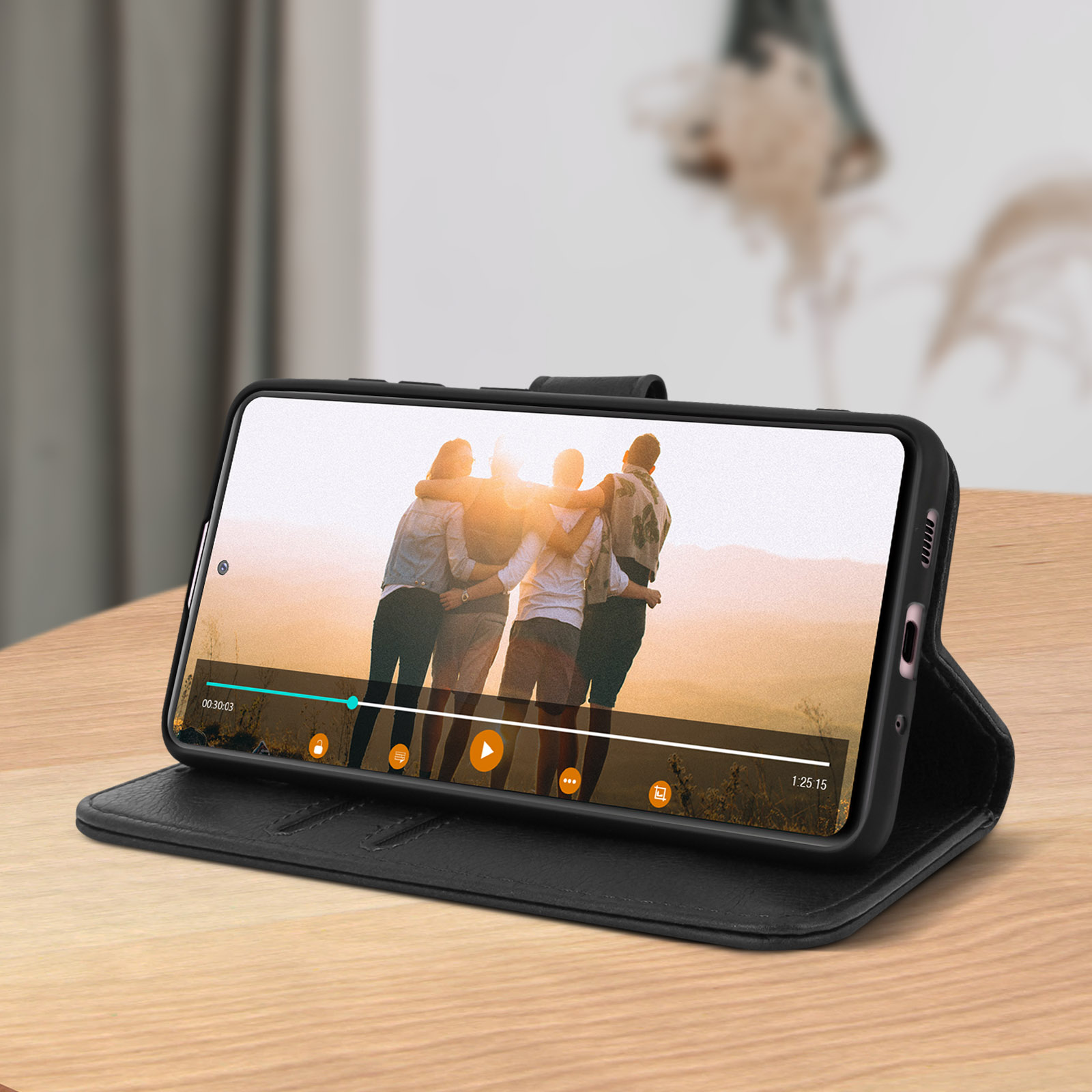 Téléphone dans un étui noir, lisant une vidéo. Il y a des personnes sur l'écran. Il est sur une table en bois.