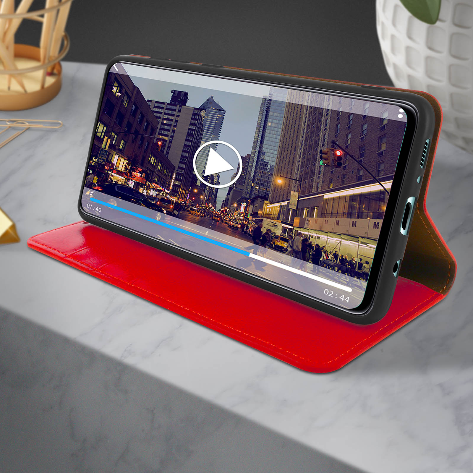 Telefono che mostra la riproduzione video. Una custodia rossa, su una superficie grigia. Sfondo con edifici cittadini.