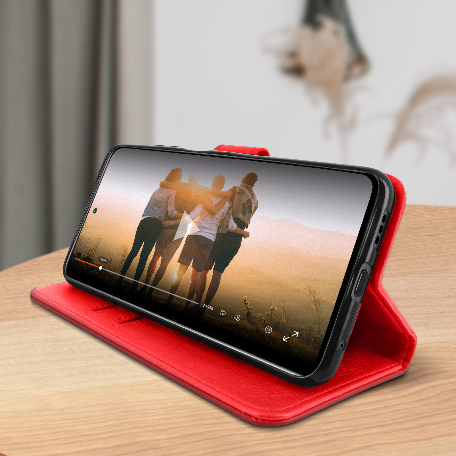 Een telefoon die een video toont van knuffelende mensen, in een rode hoes op een houten tafel. De achtergrond is wazig.