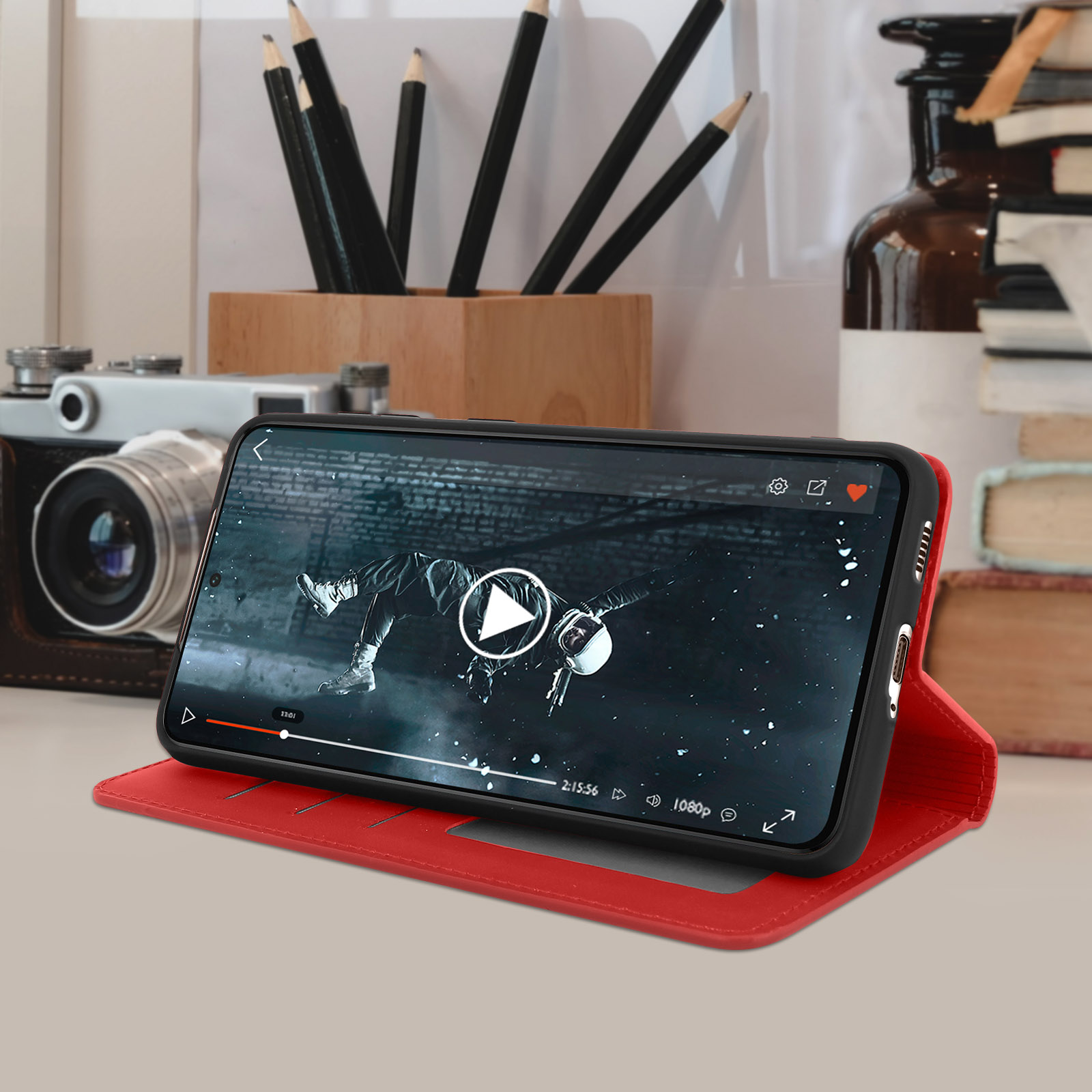 Una custodia per telefono rossa con un telefono all'interno, che riproduce un video su un tavolo con oggetti sullo sfondo.