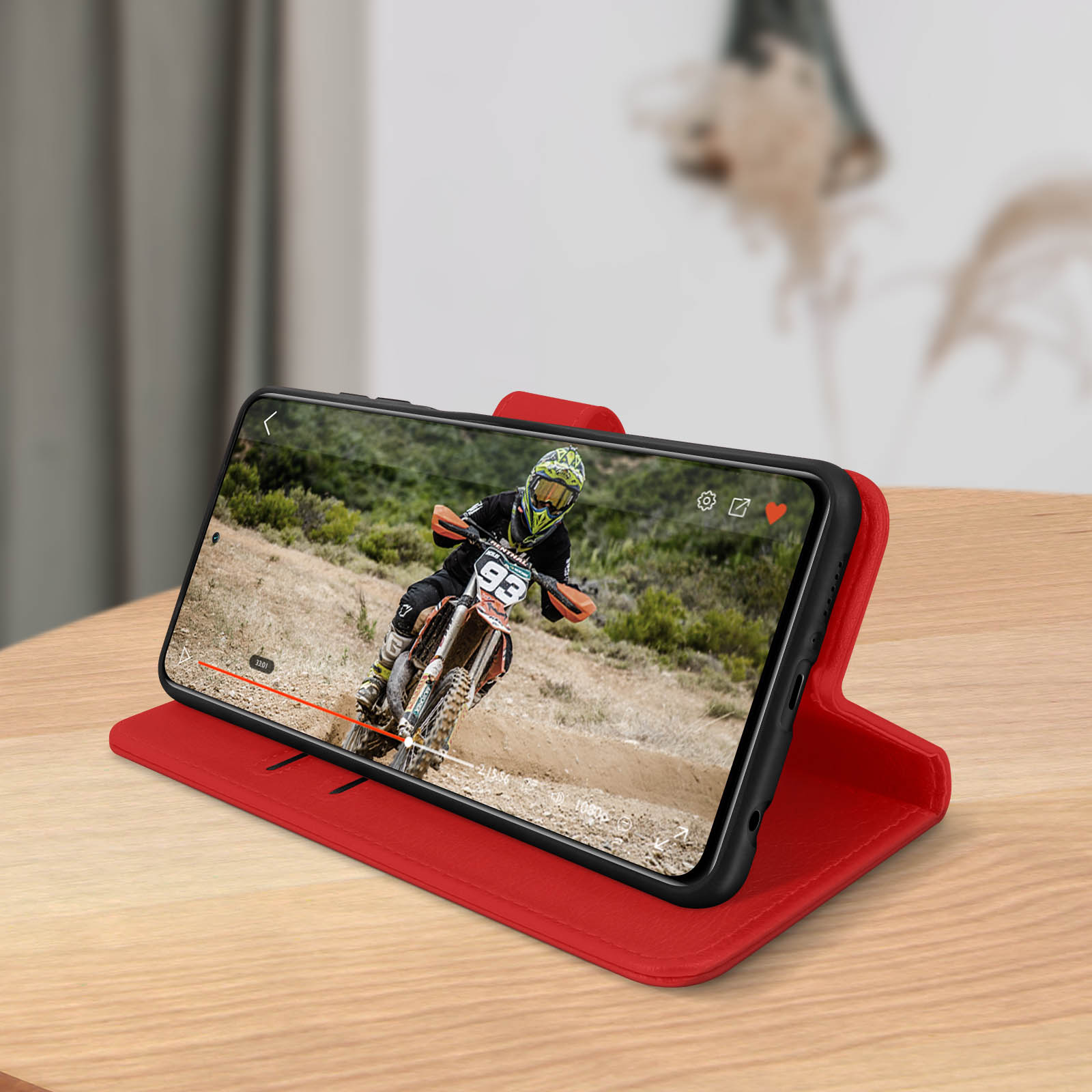 Telefono in custodia rossa, mostra un video di un motociclista su un tavolo.