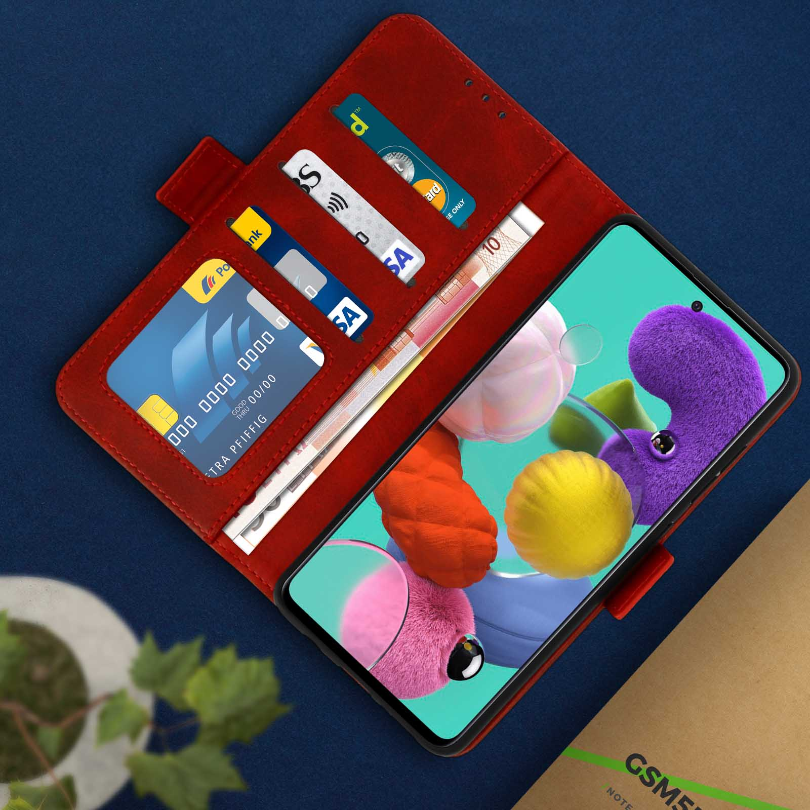 Un telefono con schermo colorato è dentro una custodia rossa aperta, accanto a carte di credito e denaro. Un quaderno è sullo sfondo.
