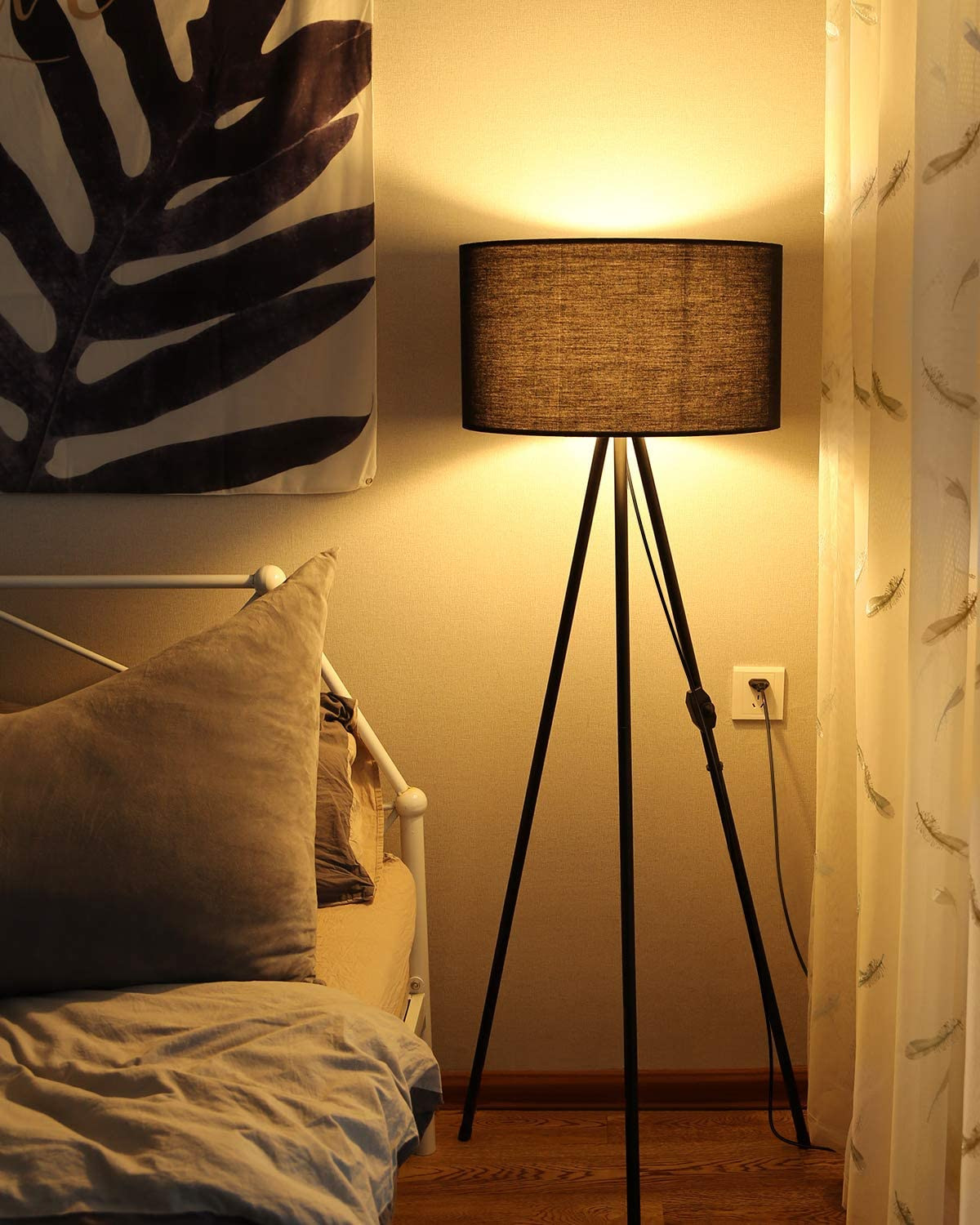 Eine beleuchtete Lampe mit schwarzem Lampenschirm auf einem Stativ. Ein Bett und Vorhänge sind im Hintergrund.