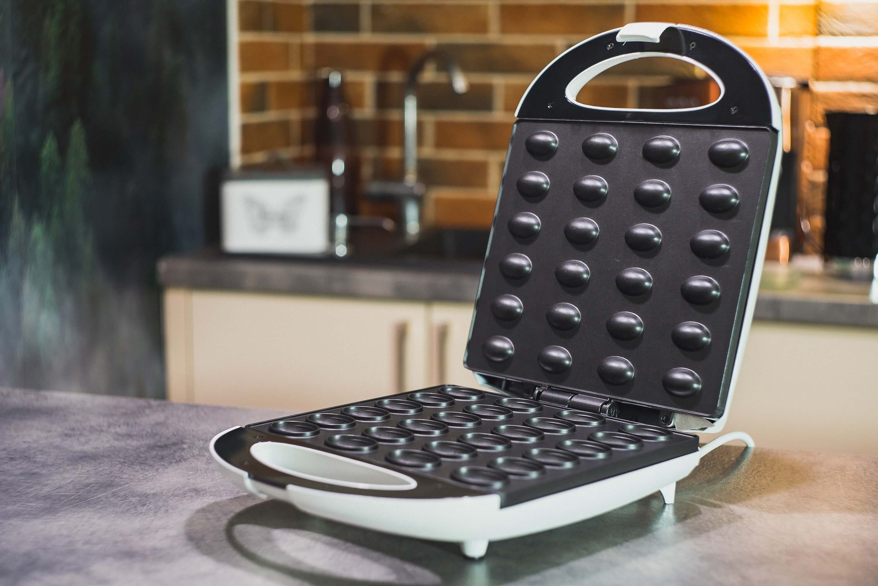 Ein Waffeleisen ist auf einer Theke geöffnet. Es hat viele kleine runde Vertiefungen. Der Hintergrund ist eine Küche.