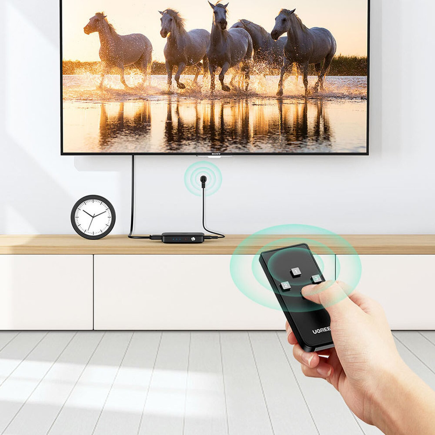 Telewizor, czarny dekoder, pilot i dłoń trzymająca pilota. Widoczny zegar i konie.