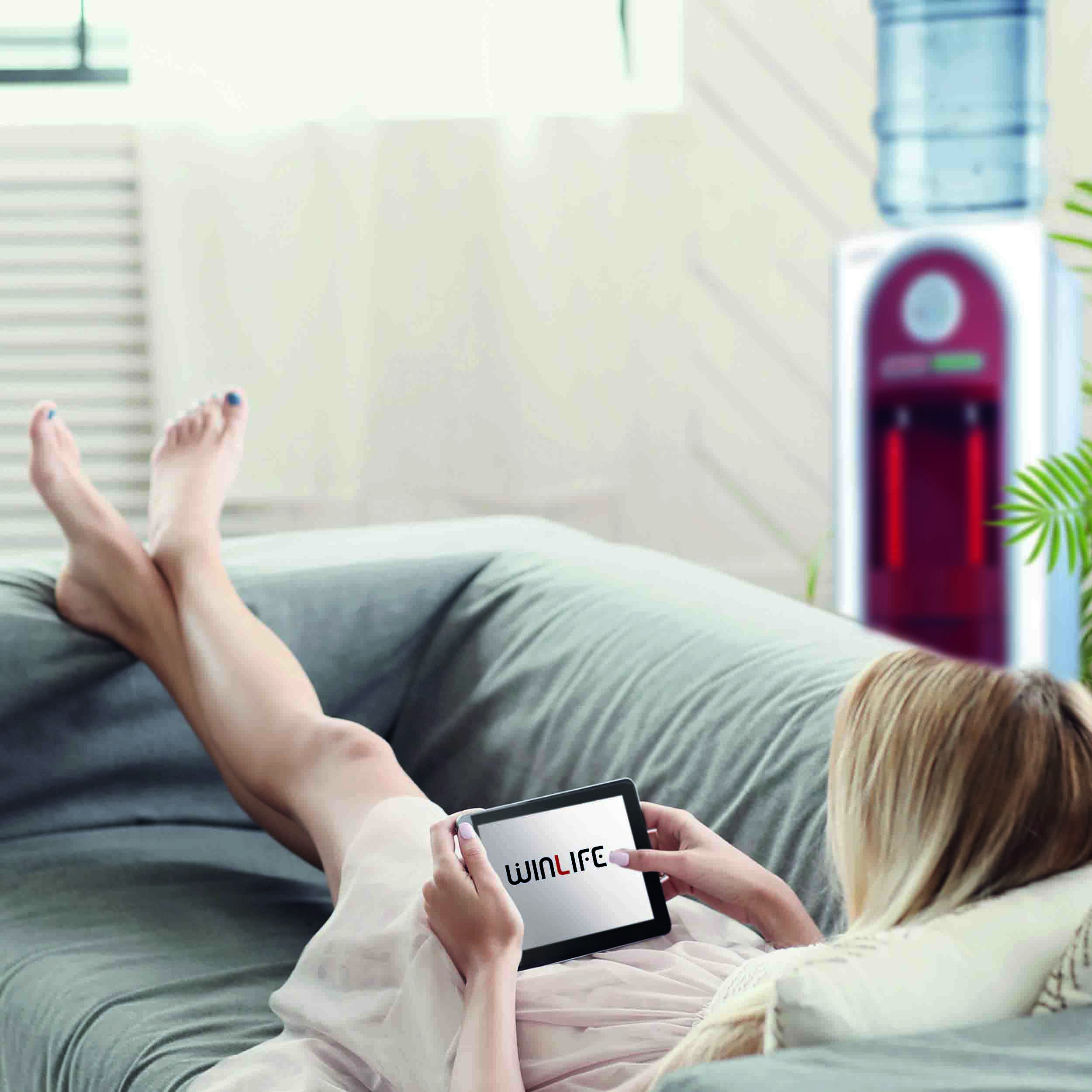 Frau auf einem grauen Sofa, die ein Tablet benutzt, mit einem Wasserspender im Hintergrund.