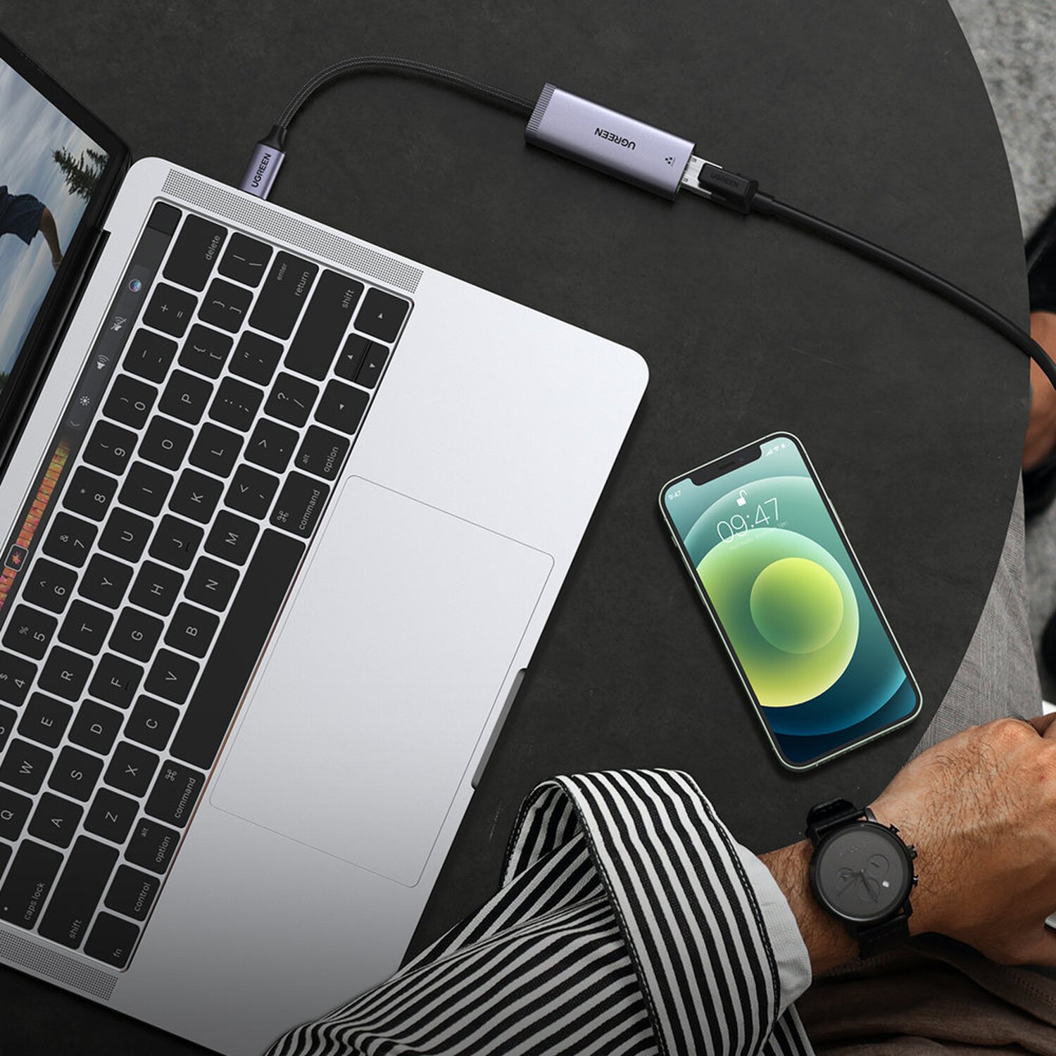 Laptop podłączony do telefonu i innego urządzenia. Osoba ma na sobie zegarek. Telefon wyświetla godzinę.