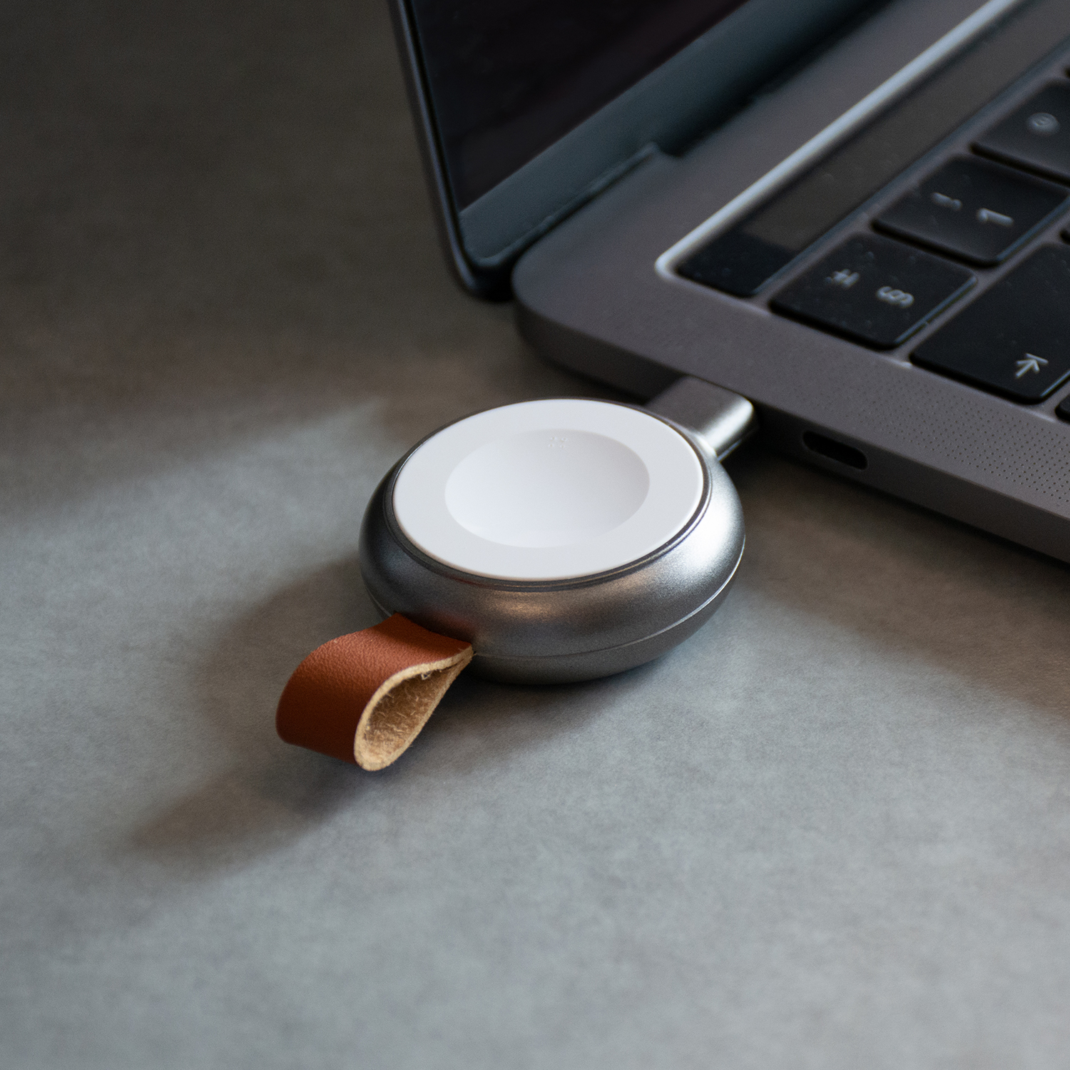 Ein silbernes Apple Watch Ladegerät, das mit einem Laptop verbunden ist. Graue Oberfläche.