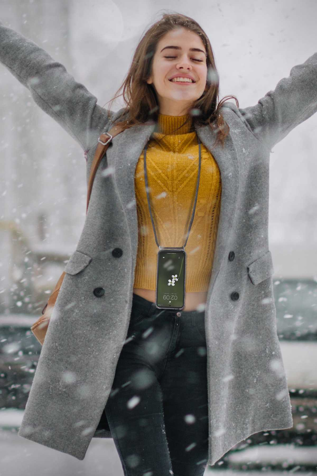 Frau in grauem Mantel, die ein Telefon mit einem Schlüsselband trägt, lächelnd, Schnee im Hintergrund.