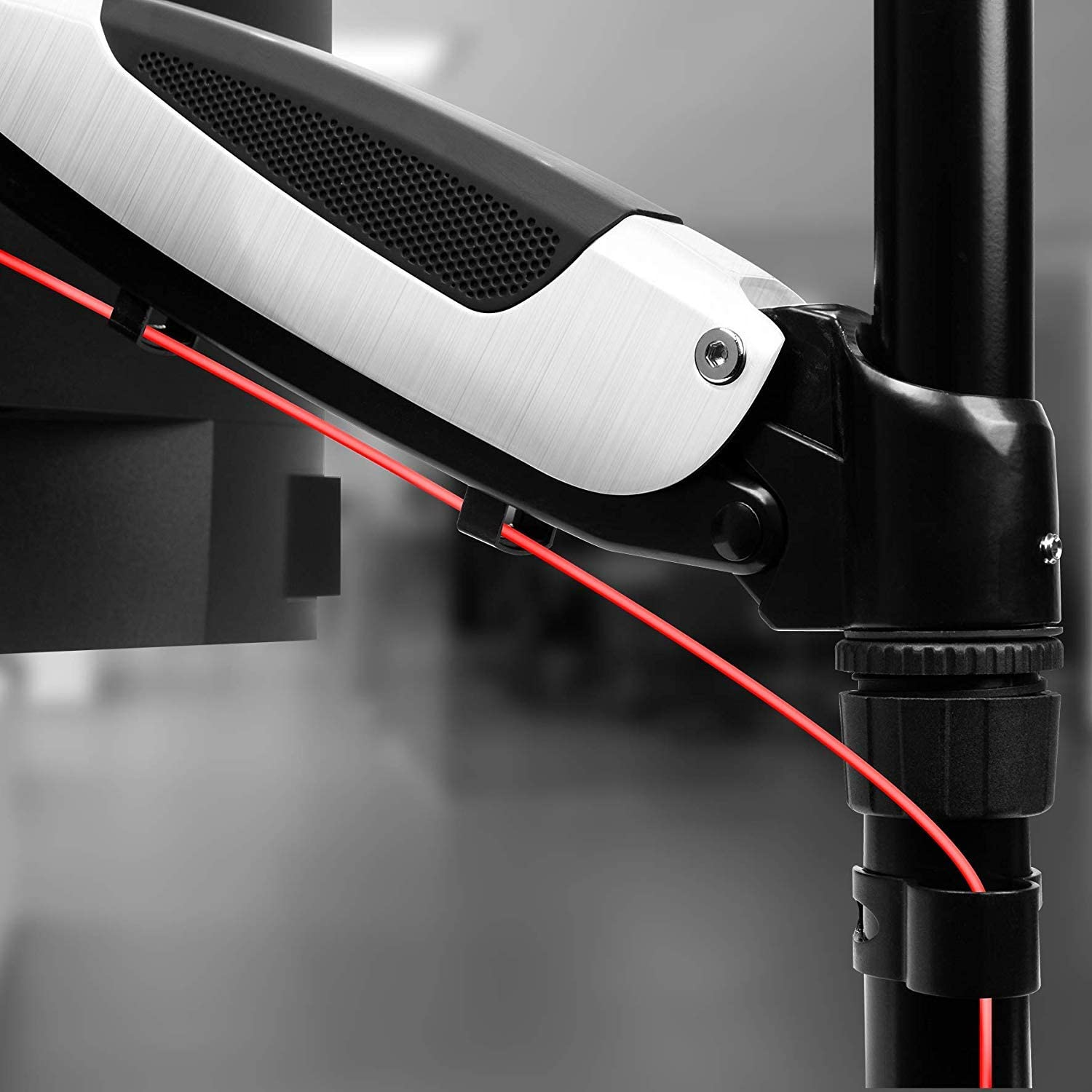Brazo de monitor con cable rojo, partes negras y plateadas. Fondo borroso.