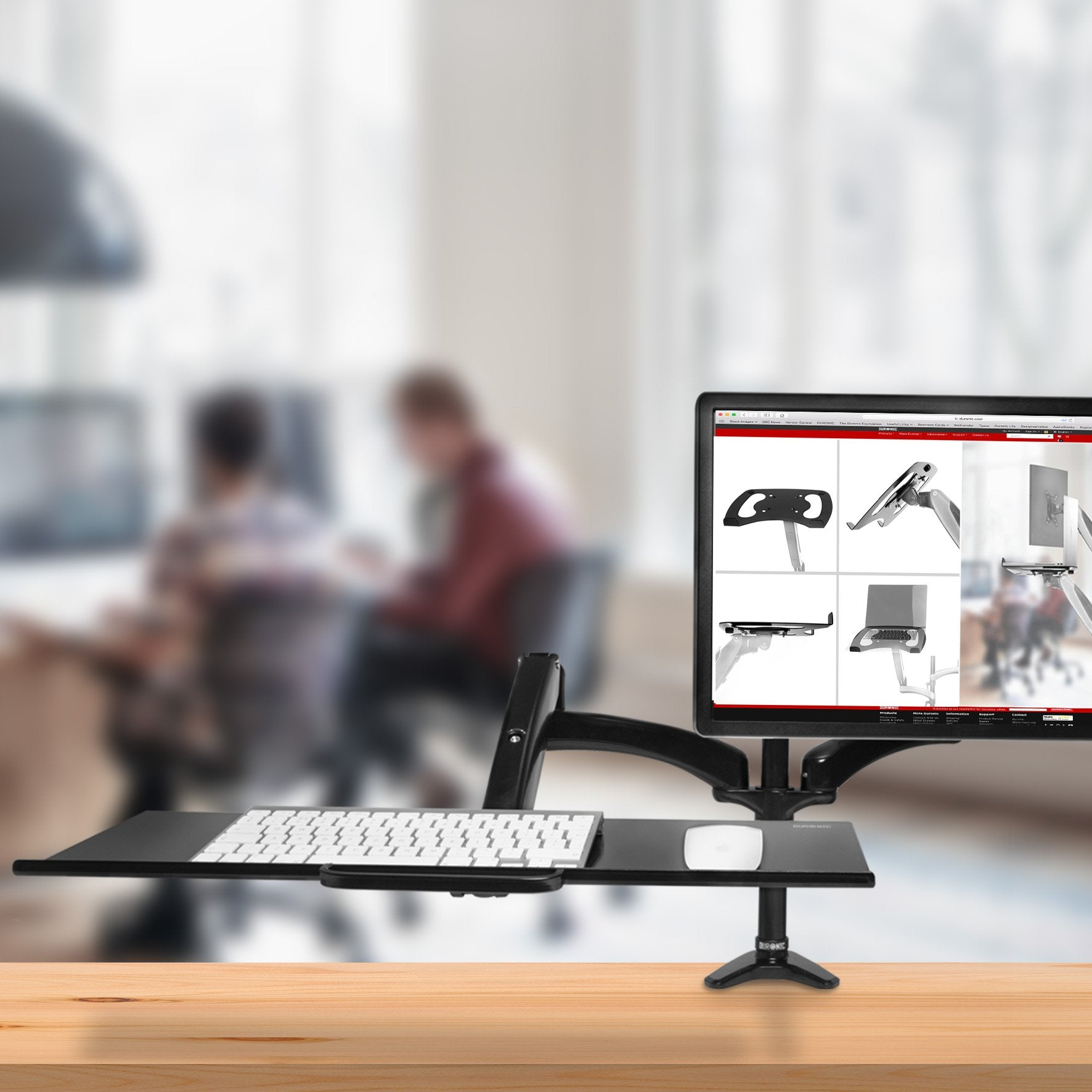 Configuración de brazo de monitor con teclado y ratón. Dos personas borrosas en el fondo.