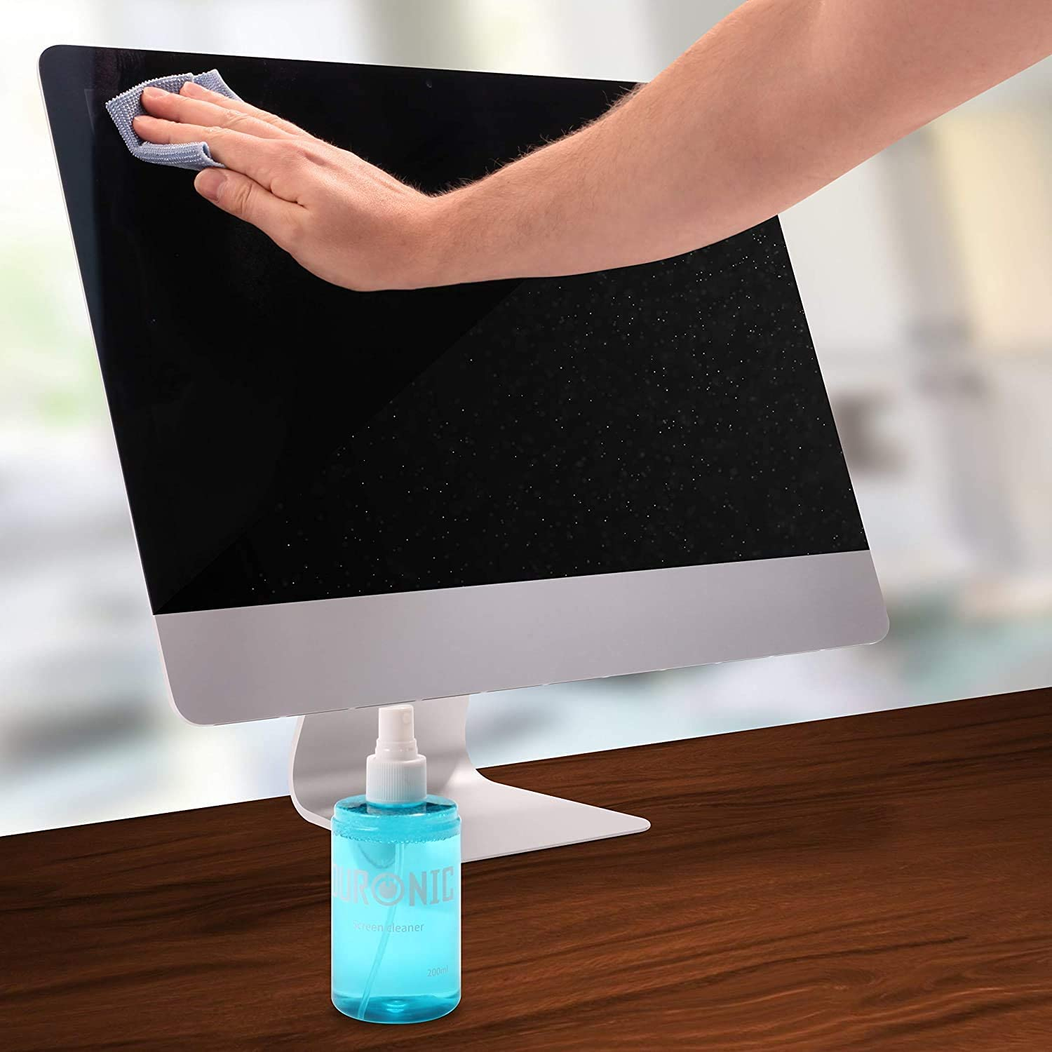 Eine Hand wischt einen Computerbildschirm mit einem Tuch ab. Eine Reinigungs-Sprühflasche steht auf dem Holztisch.