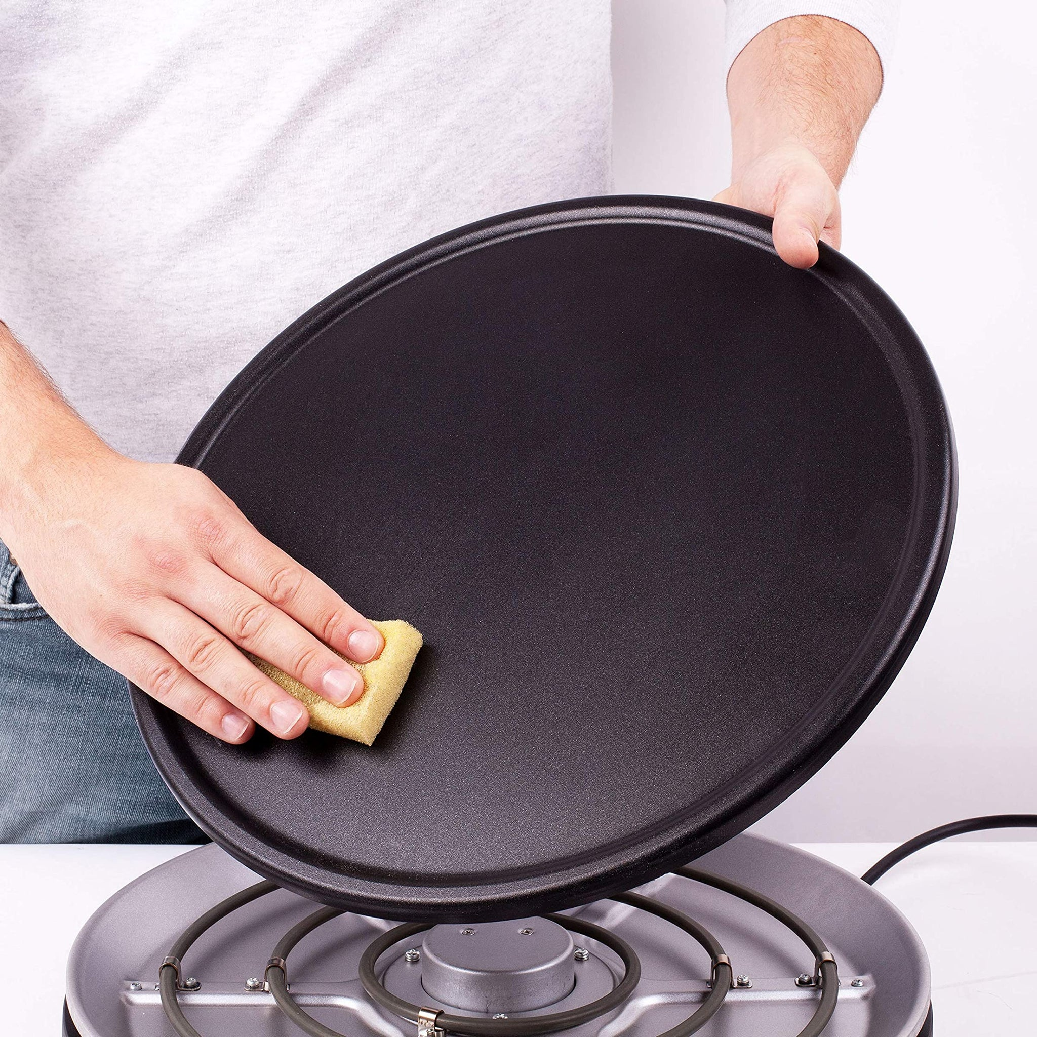 Eine Person reinigt eine schwarze Bratpfanne mit einem gelben Schwamm auf einem grauen Elektroherd.
