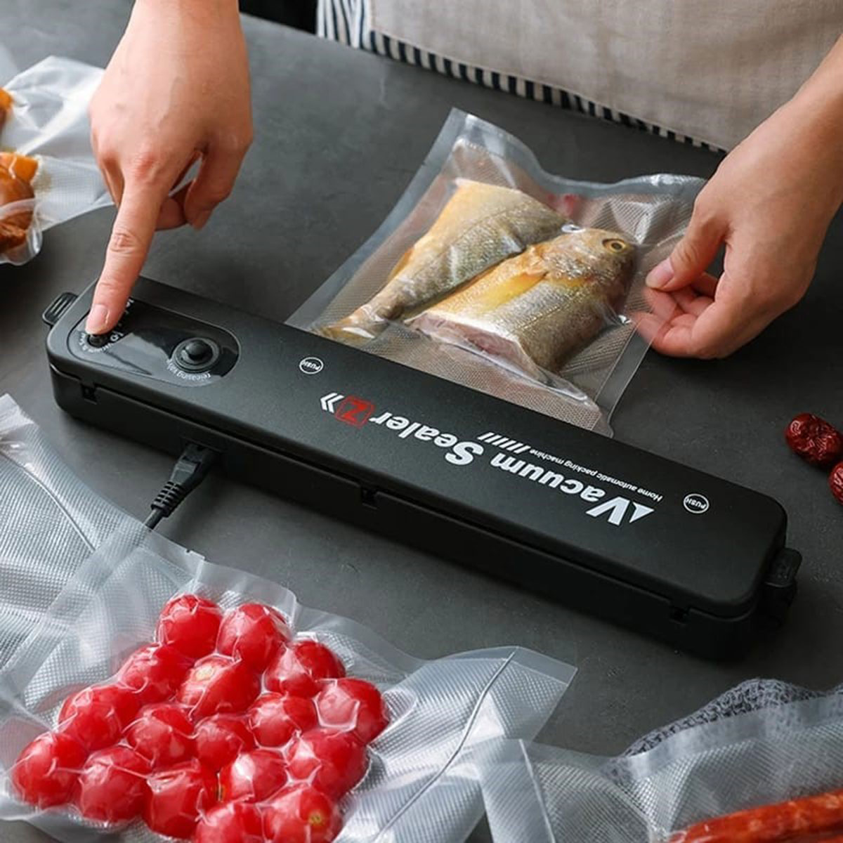 Una selladora al vacío negra con tomates y pescado en bolsas de plástico. Una persona está presionando el botón.