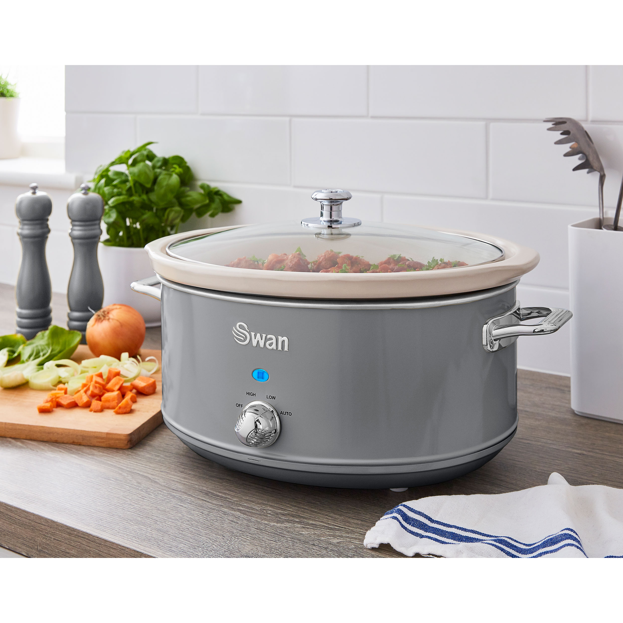 Ein grauer Slow Cooker mit Glasdeckel, Gemüse und Kräutern auf einer Küchentheke.